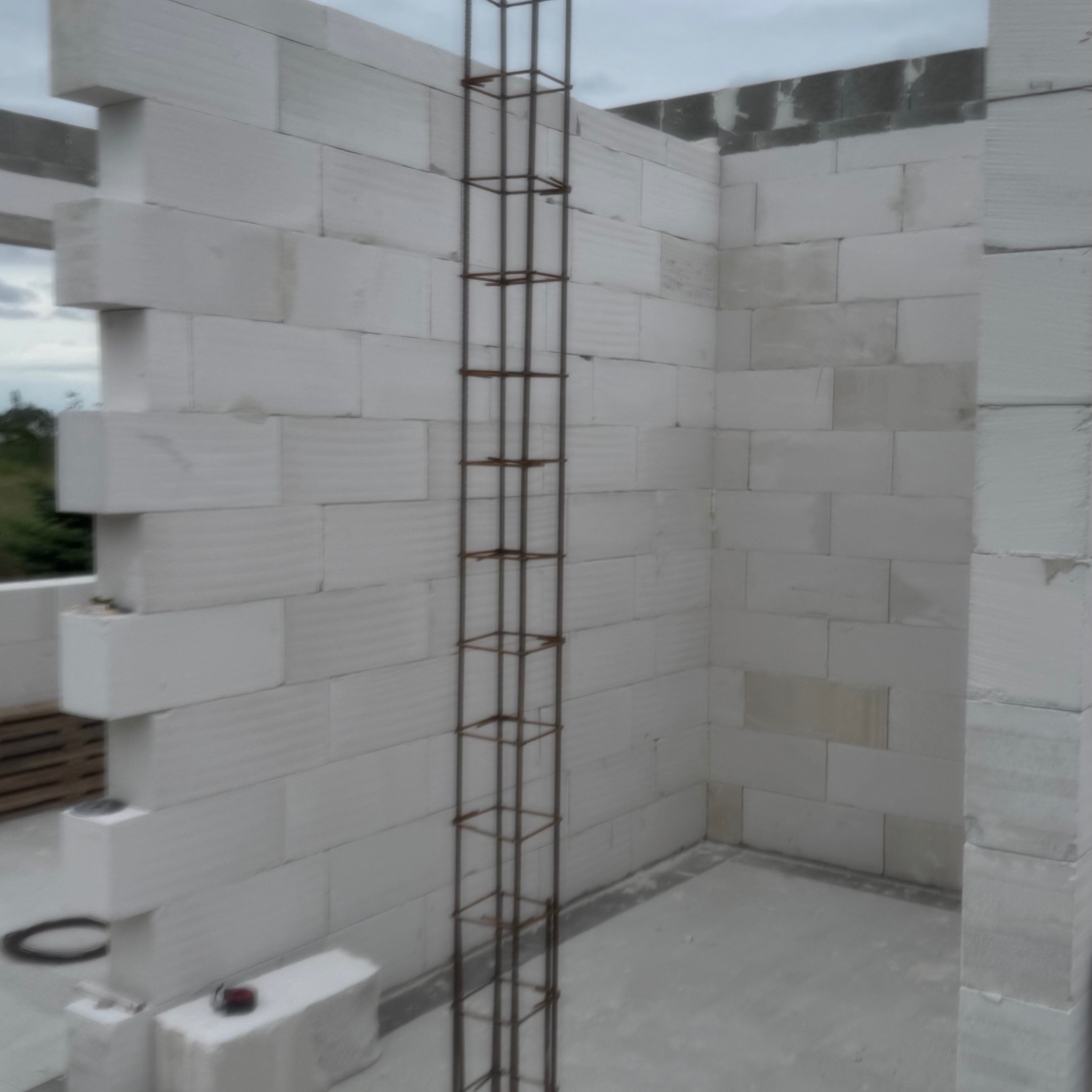 Wznoszenie ścian z białych bloczków gazobetonowych z pionowym zbrojeniem w narożniku. Widoczne narzędzia pomiarowe. Surowy stan budowy domu.