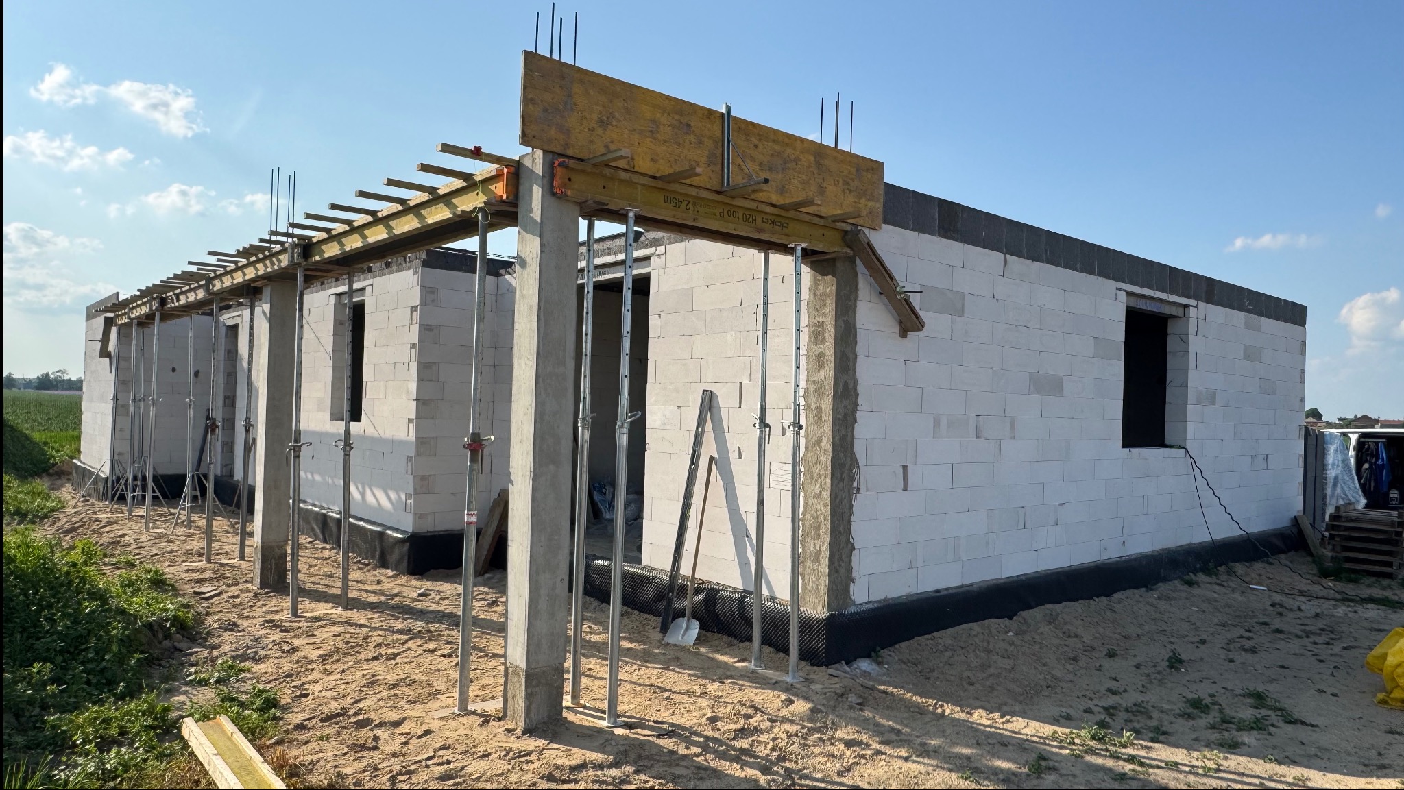 Wznoszenie ścian domu z betonu komórkowego, widoczne podpory szalunkowe i zbrojenie. Etap budowy w słoneczny dzień, zarys pól w tle.