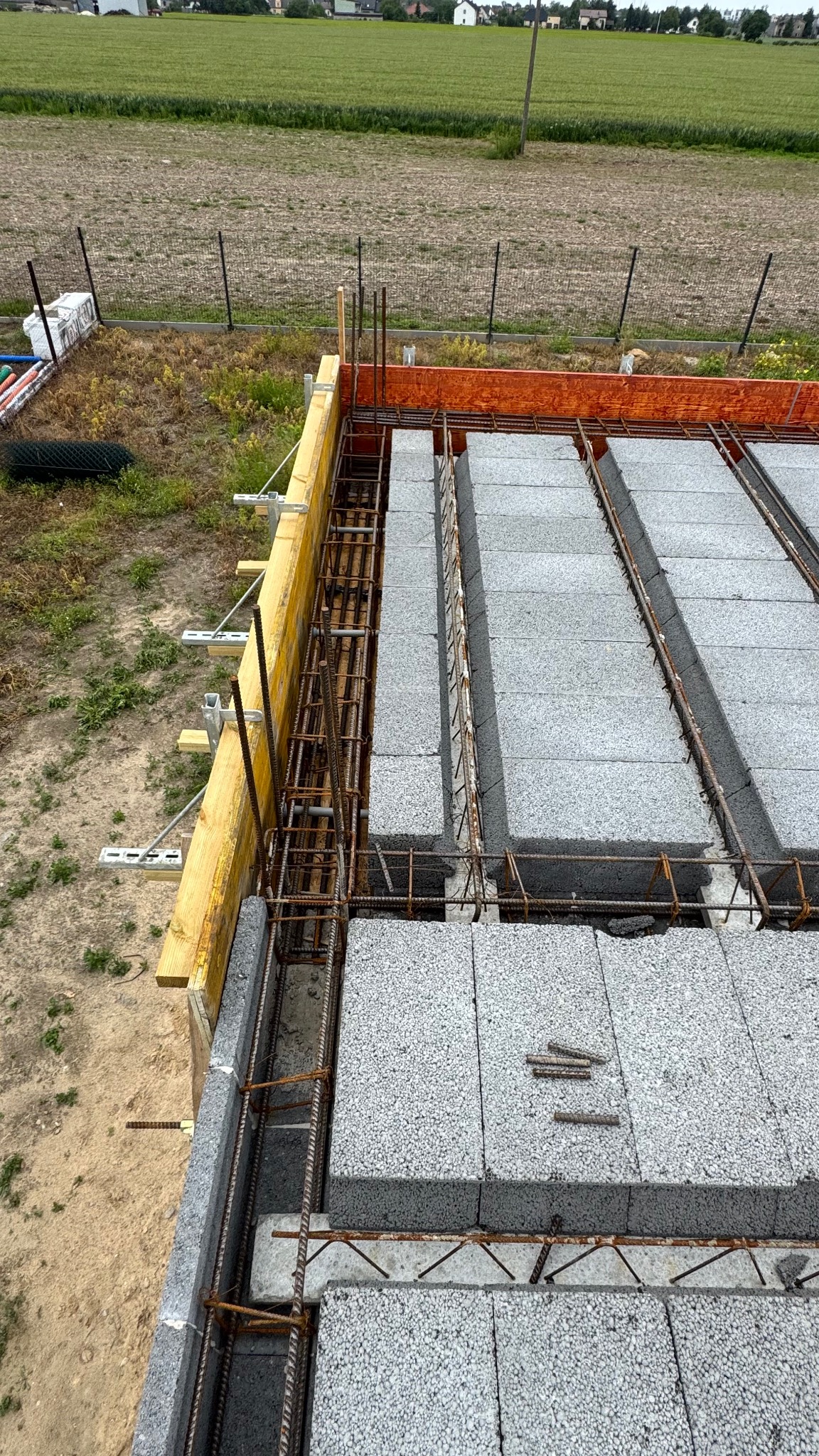 Budowa stropu Teriva: szalunek z desek, zbrojenie i pustaki stropowe. Widok z góry na plac budowy z zielonym polem w tle. Elementy konstrukcyjne w surowym stanie.