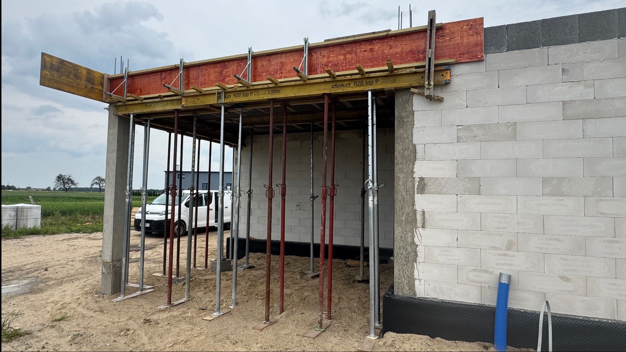 Budowa domu z betonu komórkowego: ściany wzniesione, widoczne podpory szalunkowe, szalunek stropu. Na zewnątrz zaparkowany biały samochód dostawczy.