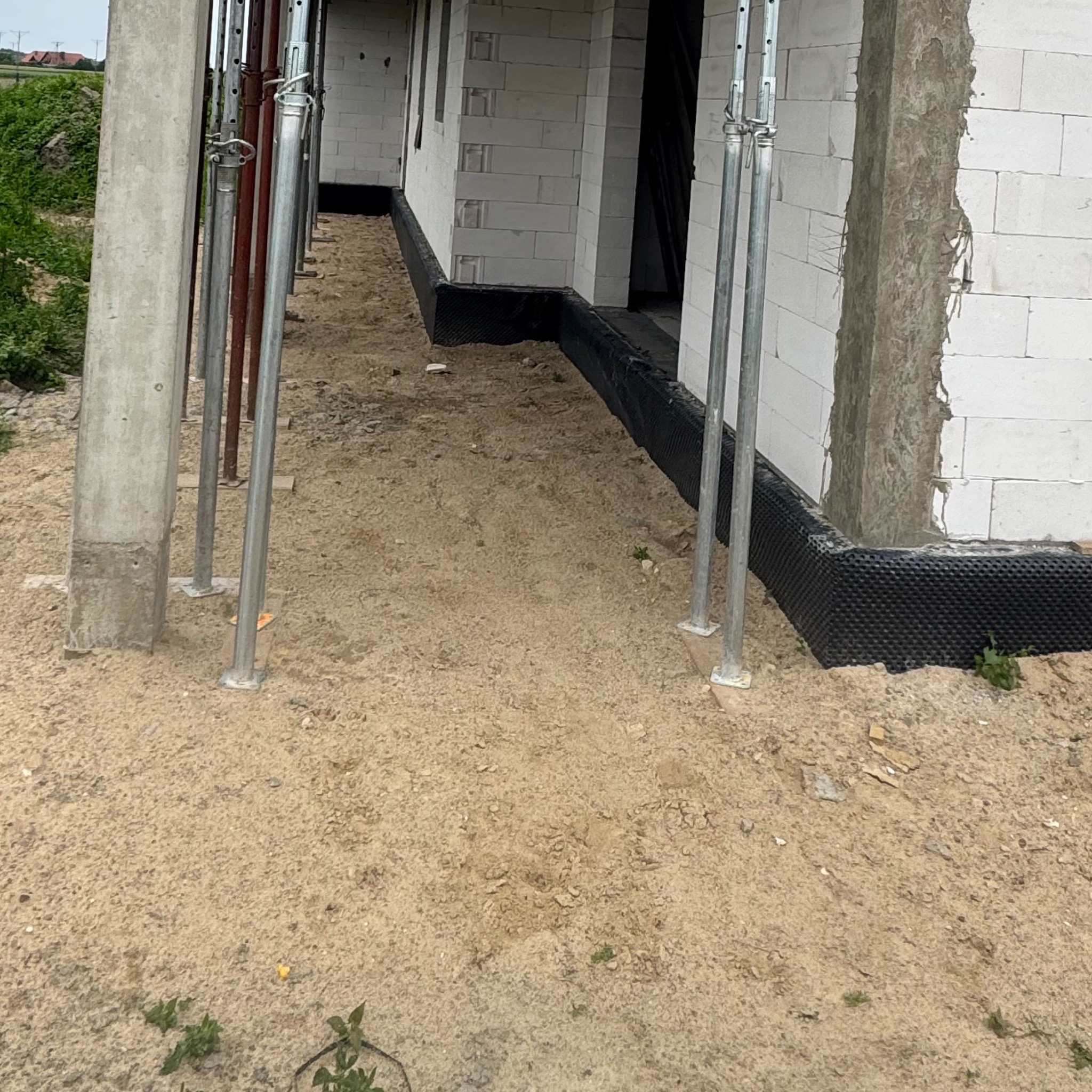 Wznoszenie ścian z betonu komórkowego na fundamencie z izolacją przeciwwilgociową. Widoczne podpory i piasek na gruncie.