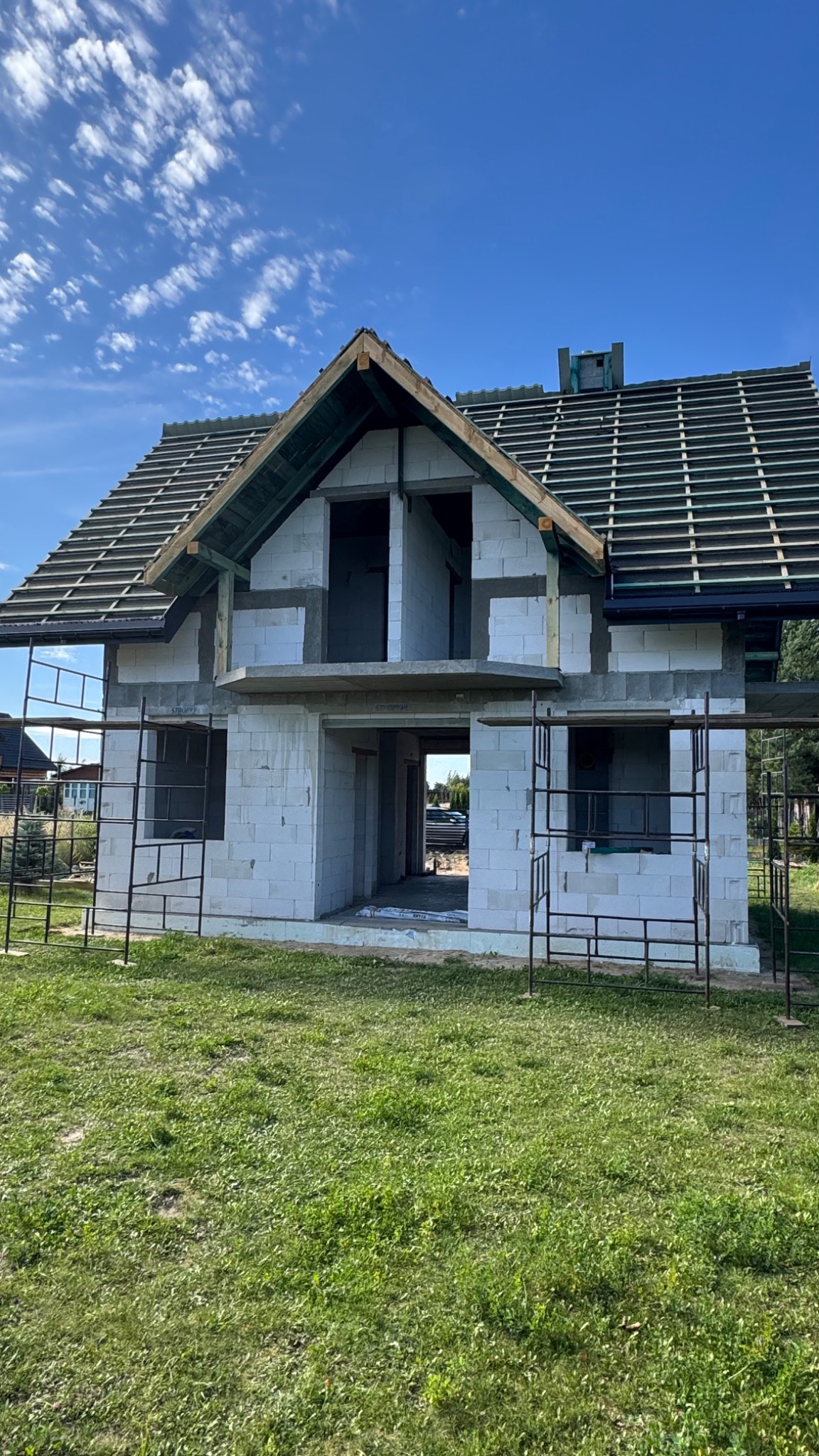 Surowy dom z białego pustaka w trakcie budowy, z częściowo położonym dachem i widocznymi rusztowaniami na tle błękitnego nieba z chmurami.