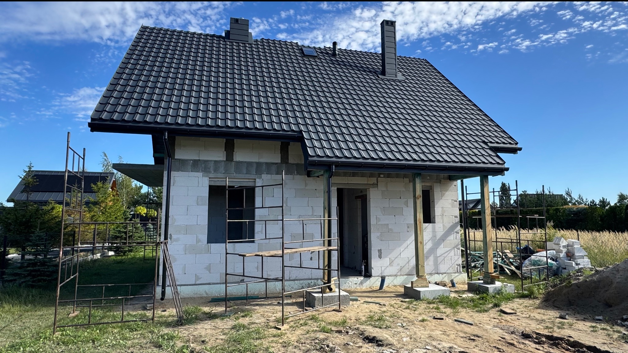Niedokończony dom z białego pustaka z czarnym dachem z dachówki, widoczne rusztowania i materiały budowlane. Błękitne niebo w tle.