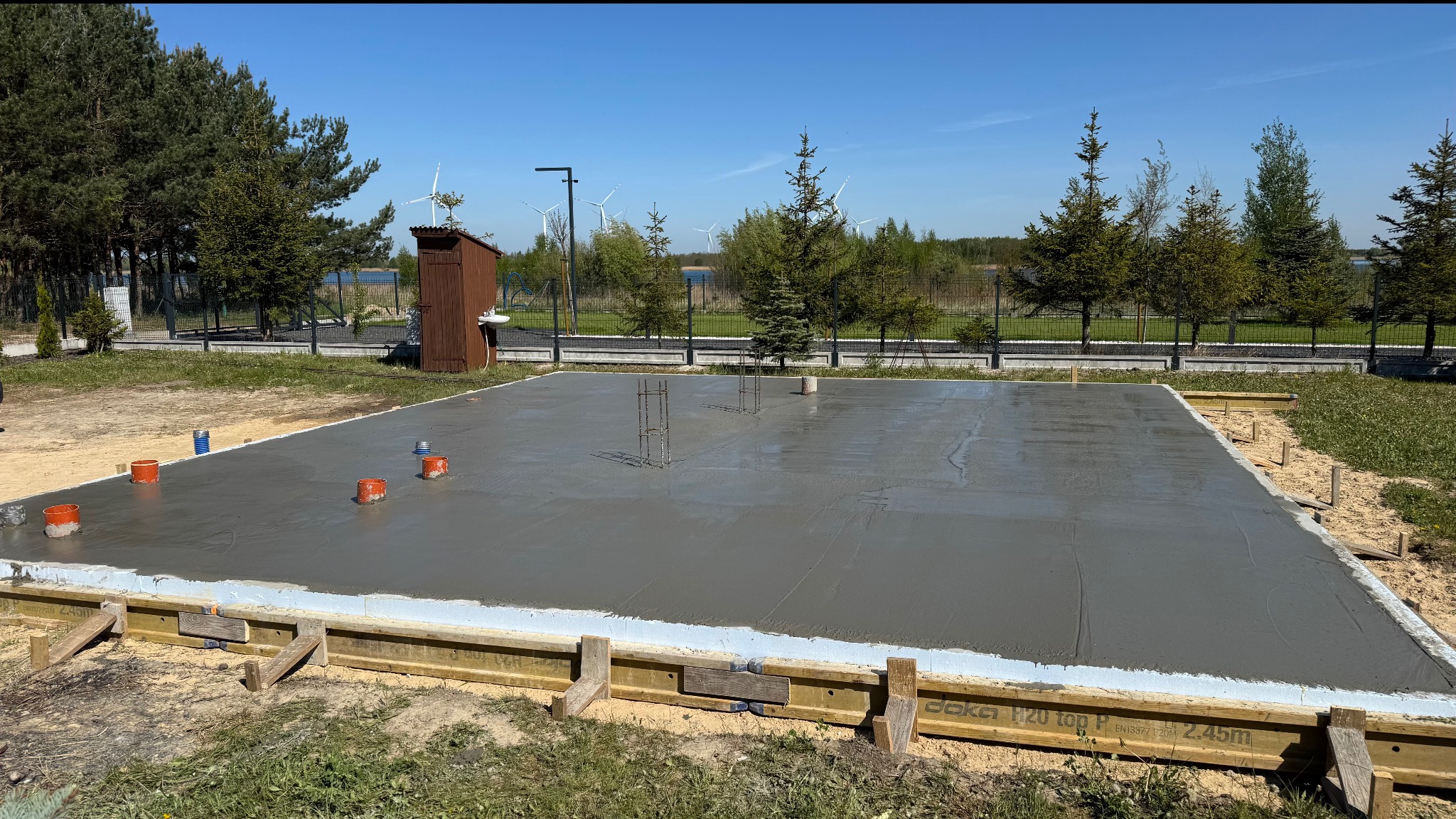 Wylana płyta fundamentowa z wystającymi prętami zbrojeniowymi i elementami instalacji, otoczona drewnianym szalunkiem na tle zieleni i błękitnego nieba.