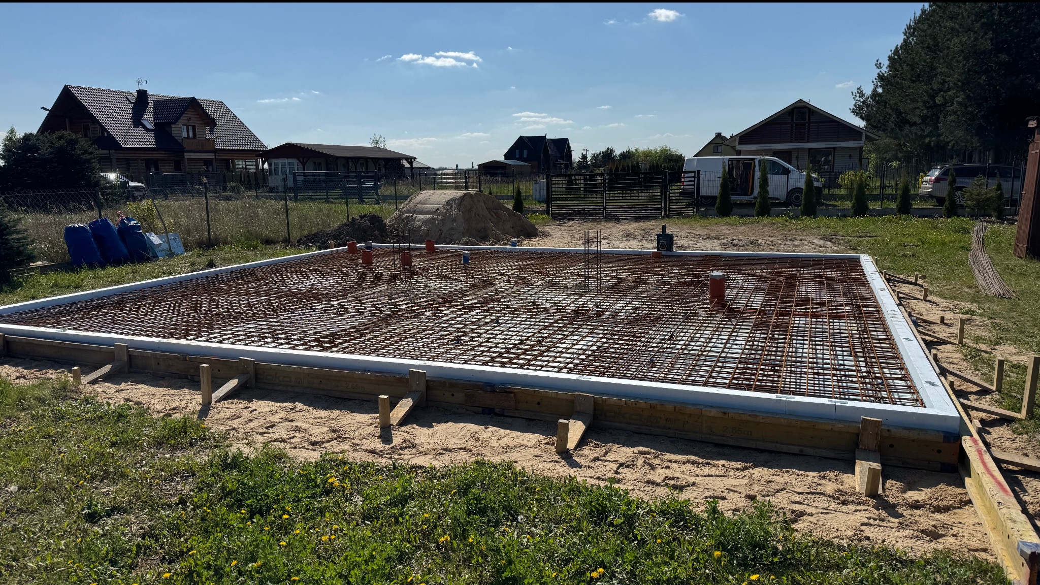 Zbrojenie fundamentu domu jednorodzinnego z widocznymi elementami instalacji, otoczone szalunkiem i ociepleniem, na tle zabudowy jednorodzinnej w słoneczny dzień.