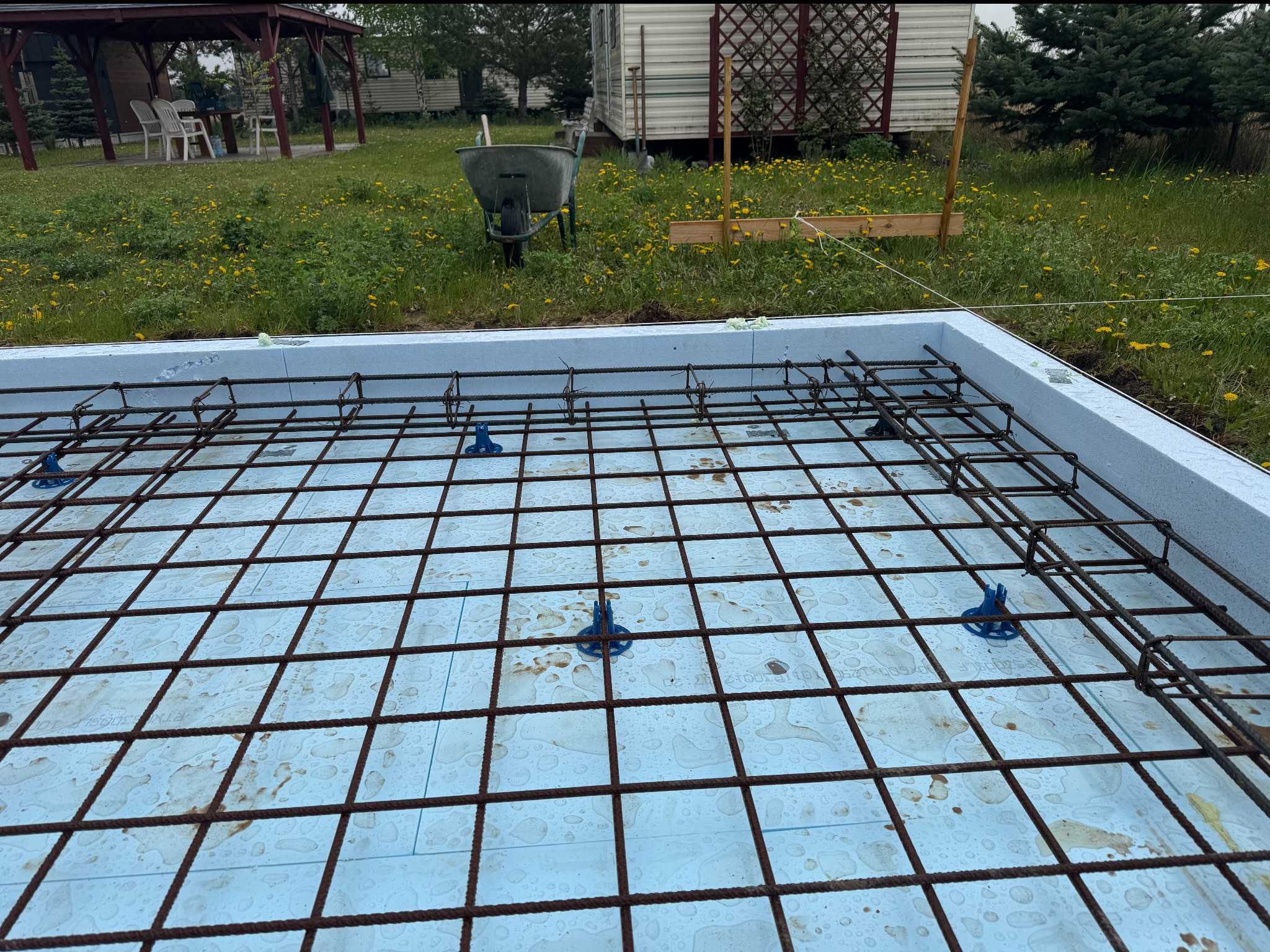 Zbrojenie fundamentu na styropianie z widocznymi podkładkami dystansowymi, przygotowane do zalania betonem. W tle taczka i teren budowy.