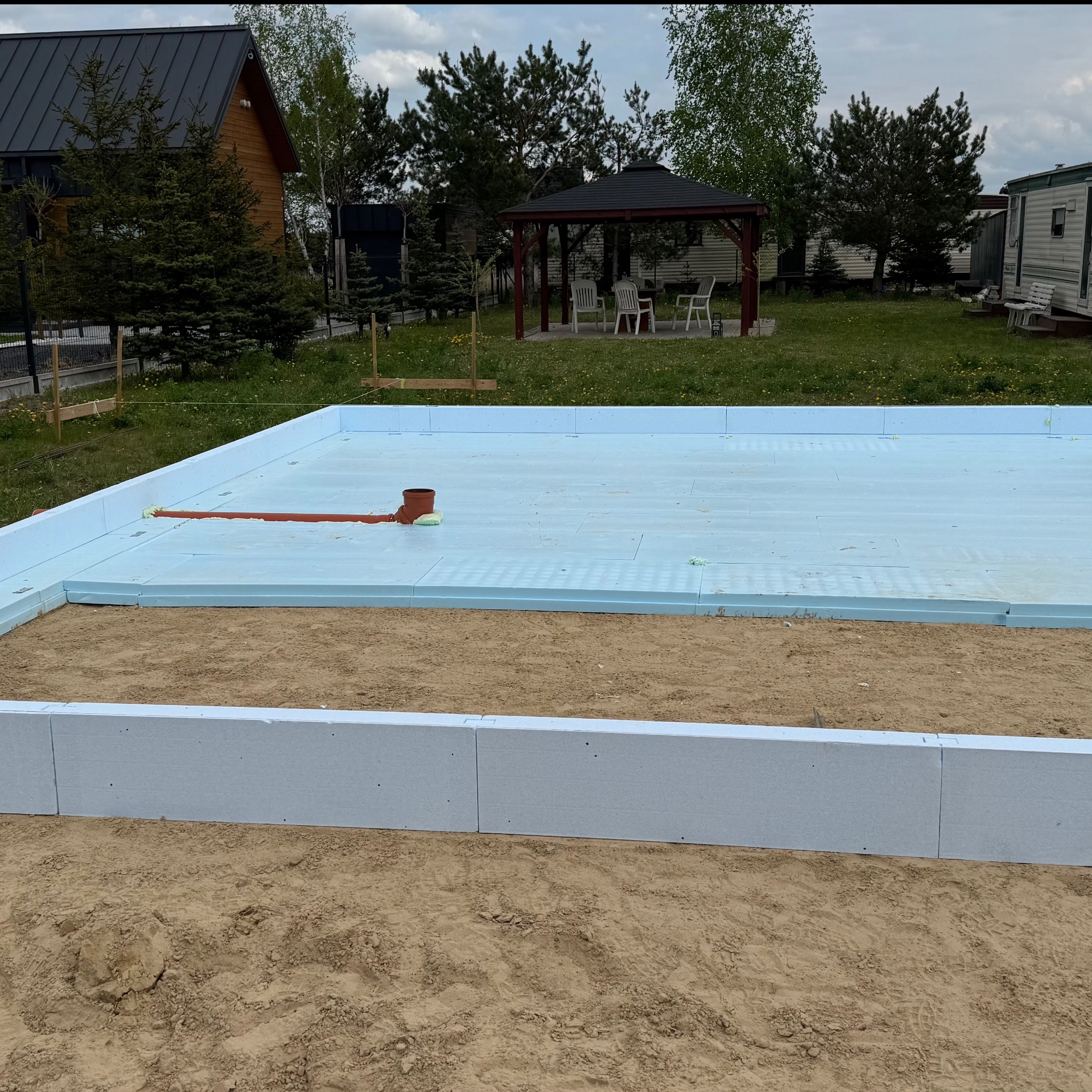 Izolacja fundamentów domu jednorodzinnego z widoczną instalacją kanalizacyjną, przygotowana do dalszych prac budowlanych na działce z altaną i domkami letniskowymi w tle.