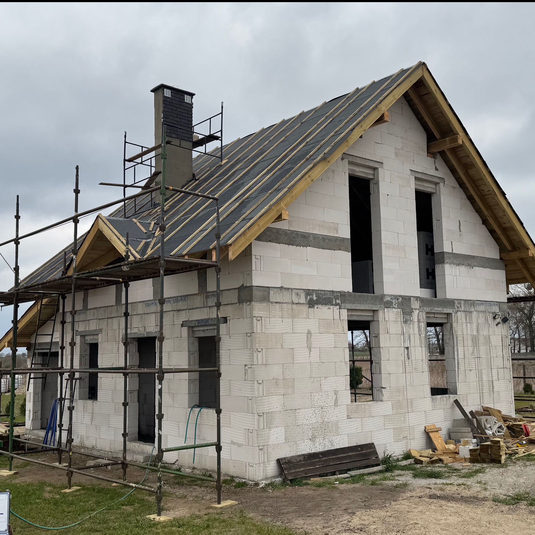 Surowy dom z białego pustaka w trakcie budowy, z widocznym dachem pokrytym papą i rusztowaniami. Ujęcie z zewnątrz, w pochmurny dzień.