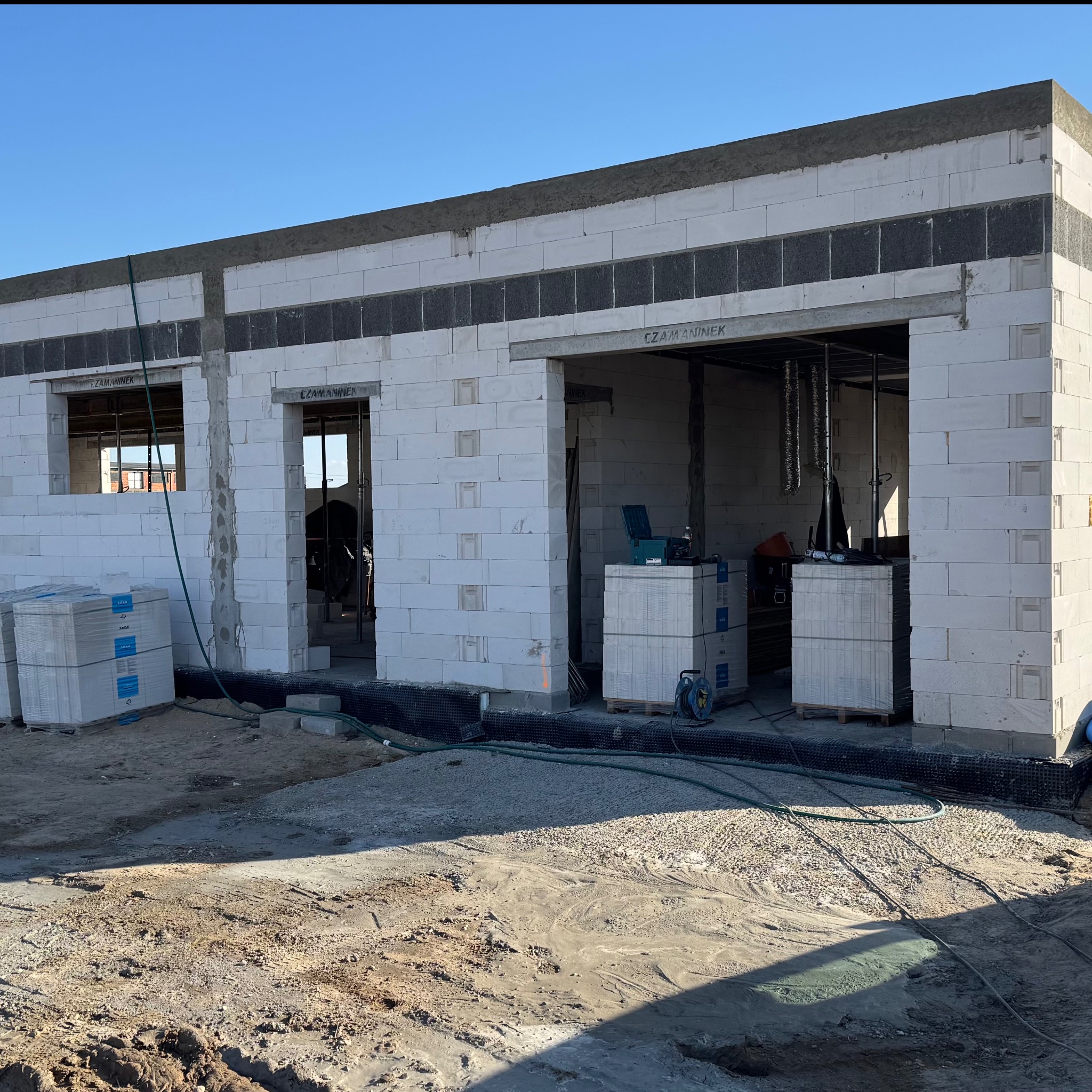 Surowy stan budynku z białych bloczków, widoczne otwory okienne i drzwiowe, palety z materiałami budowlanymi, teren budowy w słoneczny dzień.