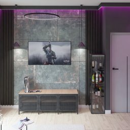 Jagoda Nowak Pracownia Projektowa - Nowoczesny salon z szaro-zieloną ścianą akcentową, podświetleniem LED, telewizorem z grą Wiedźmin, konsolą PlayStation 5, szafką RTV z metalowymi drzwiczkami, barkiem z winami oraz zasłonami.