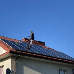 Pracownik w uprzęży i kasku myje panele słoneczne na dachu domu jednorodzinnego za pomocą szczotki na wysięgniku, na tle błękitnego nieba.