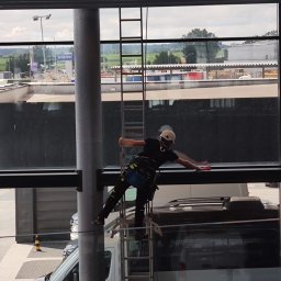 czystepanele.pl - mycie okien, usługi alpinistyczne Konrad Kusajda - Pracownik w kasku i uprzęży, na drabinie, myje okna budynku biurowego. Widok na parking z samochodami i słup z logo Volvo w tle. Czyste okno w górnej części kadru.