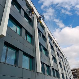 czystepanele.pl - mycie okien, usługi alpinistyczne Konrad Kusajda - Dwóch alpinistów przemysłowych myje okna na wysokościach, zabezpieczonych linami, na tle nowoczesnego biurowca z szarymi panelami i niebieskimi oknami.