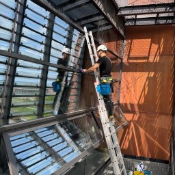 czystepanele.pl - mycie okien, usługi alpinistyczne Konrad Kusajda - Pracownik w uprzęży myje okna na wysokości, stojąc na drabinie. Widoczne refleksy nieba i otoczenia w czyszczonych oknach. Budynek o rdzawym kolorze.