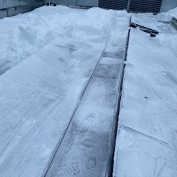 Odśnieżony fragment dachu z widocznymi śladami butów w śniegu, obok leżące pryzmy śniegu. W tle instalacja wentylacyjna pokryta śniegiem.