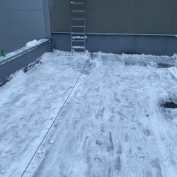 czystepanele.pl - mycie okien, usługi alpinistyczne Konrad Kusajda - Śnieg na dachu budynku po odśnieżaniu, widoczne ślady butów i lina zabezpieczająca. Metalowa drabina oparta o ścianę. Szary budynek w tle.