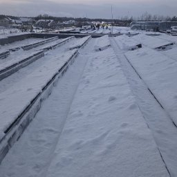 czystepanele.pl - mycie okien, usługi alpinistyczne Konrad Kusajda - Śnieg na dachu budynku z widocznymi elementami konstrukcyjnymi i pracownikami w oddali, sfotografowany w pochmurny dzień.