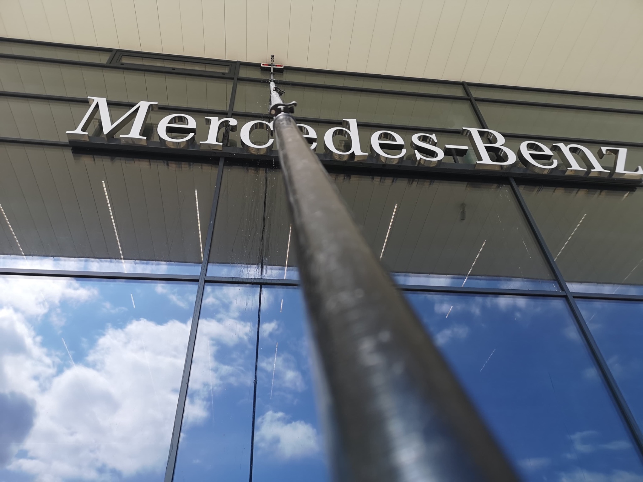 Mycie okien na wysokości budynku z napisem Mercedes-Benz. Teleskopowa lanca czyszcząca w trakcie pracy, widoczne smugi wody. Błękitne niebo odbija się w szybach.