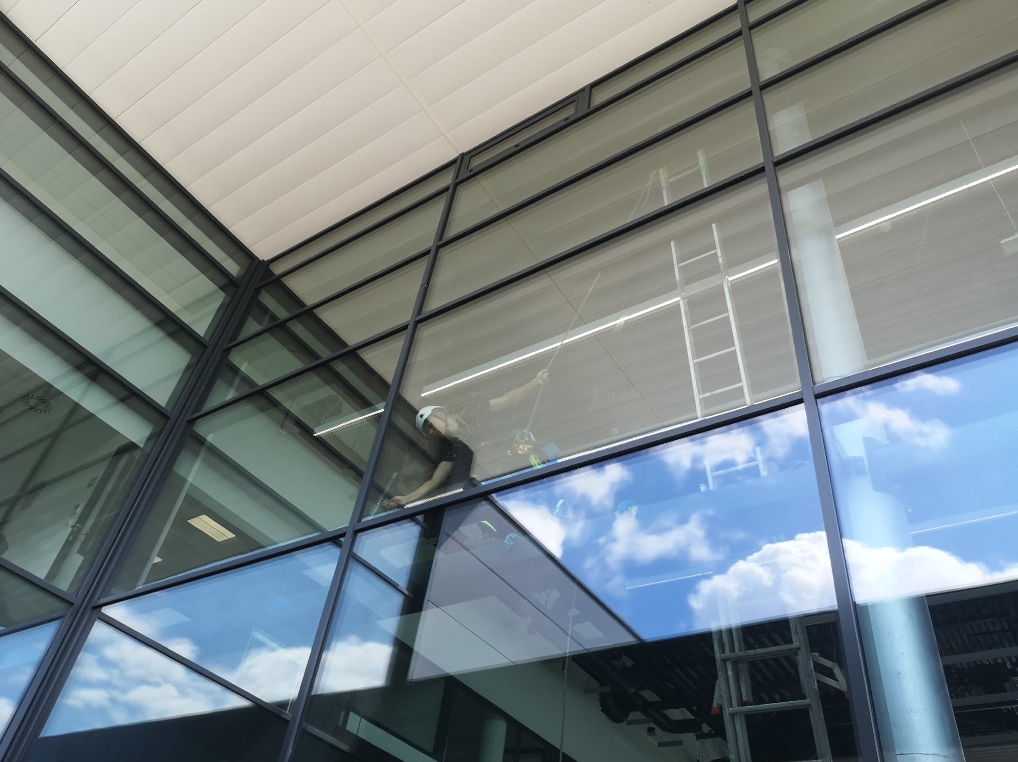 Pracownik w kasku myje okna na wysokości w nowoczesnym biurowcu z aluminiową ramą, widoczne odbicie nieba w szybach, drabina w tle.
