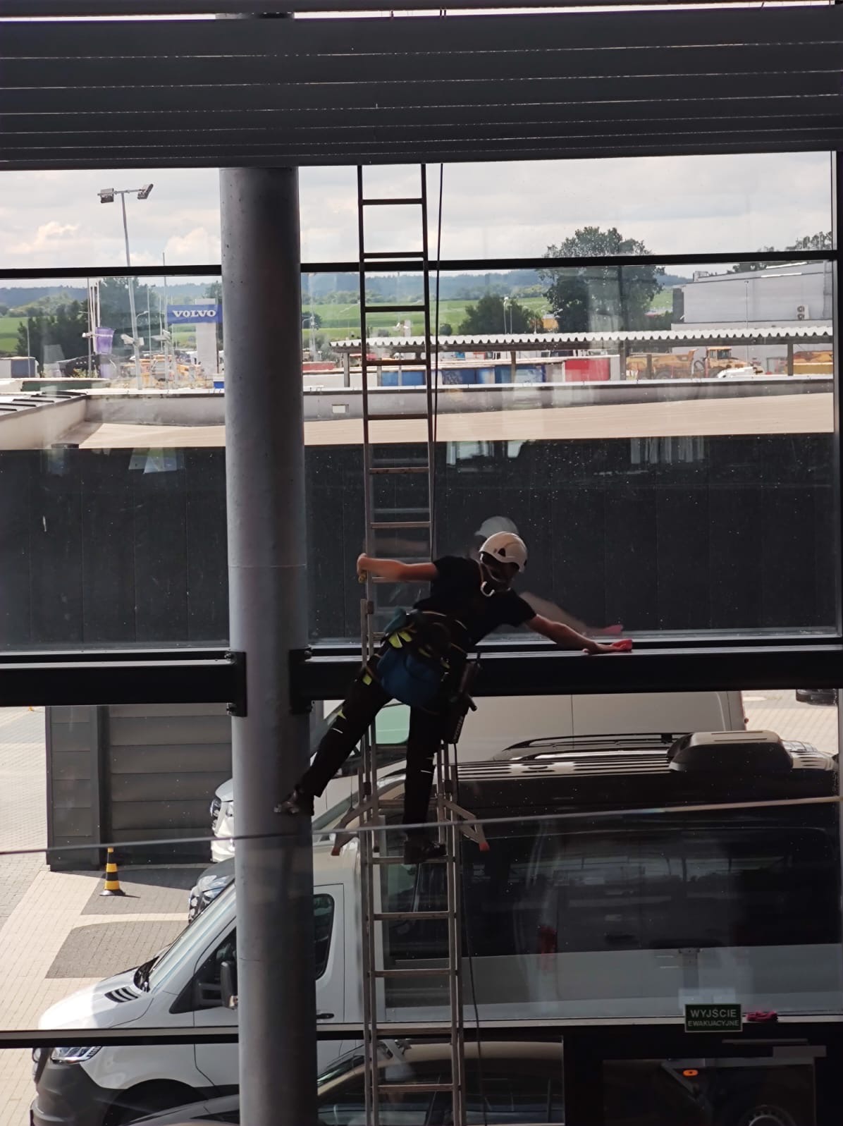 Pracownik w kasku i uprzęży, na drabinie, myje okna budynku biurowego. Widok na parking z samochodami i słup z logo Volvo w tle. Czyste okno w górnej części kadru.