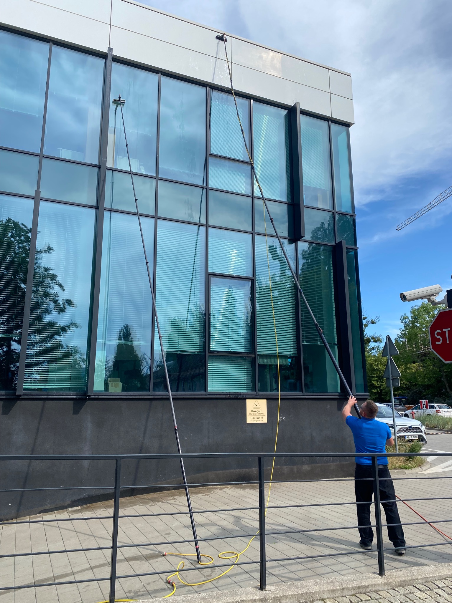 Pracownik w niebieskiej koszulce myje okna biurowca za pomocą długiej lancy teleskopowej. Widoczne odbicia w czyszczonych oknach i chodnik z kostki.