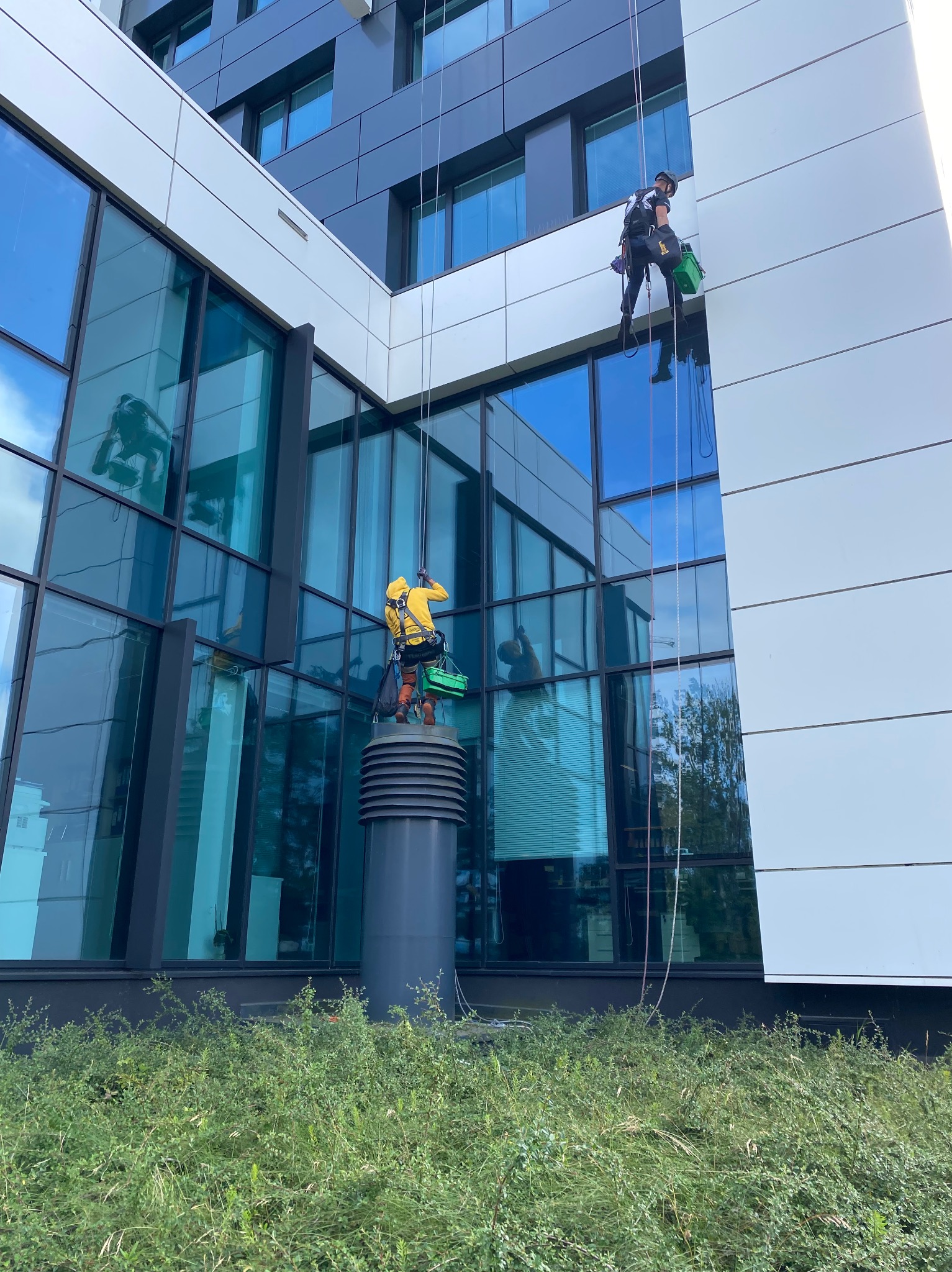 Dwóch alpinistów na linach czyści okna nowoczesnego biurowca. Jeden w żółtej kurtce stoi na rurze wentylacyjnej, drugi wyżej w czarnym stroju.