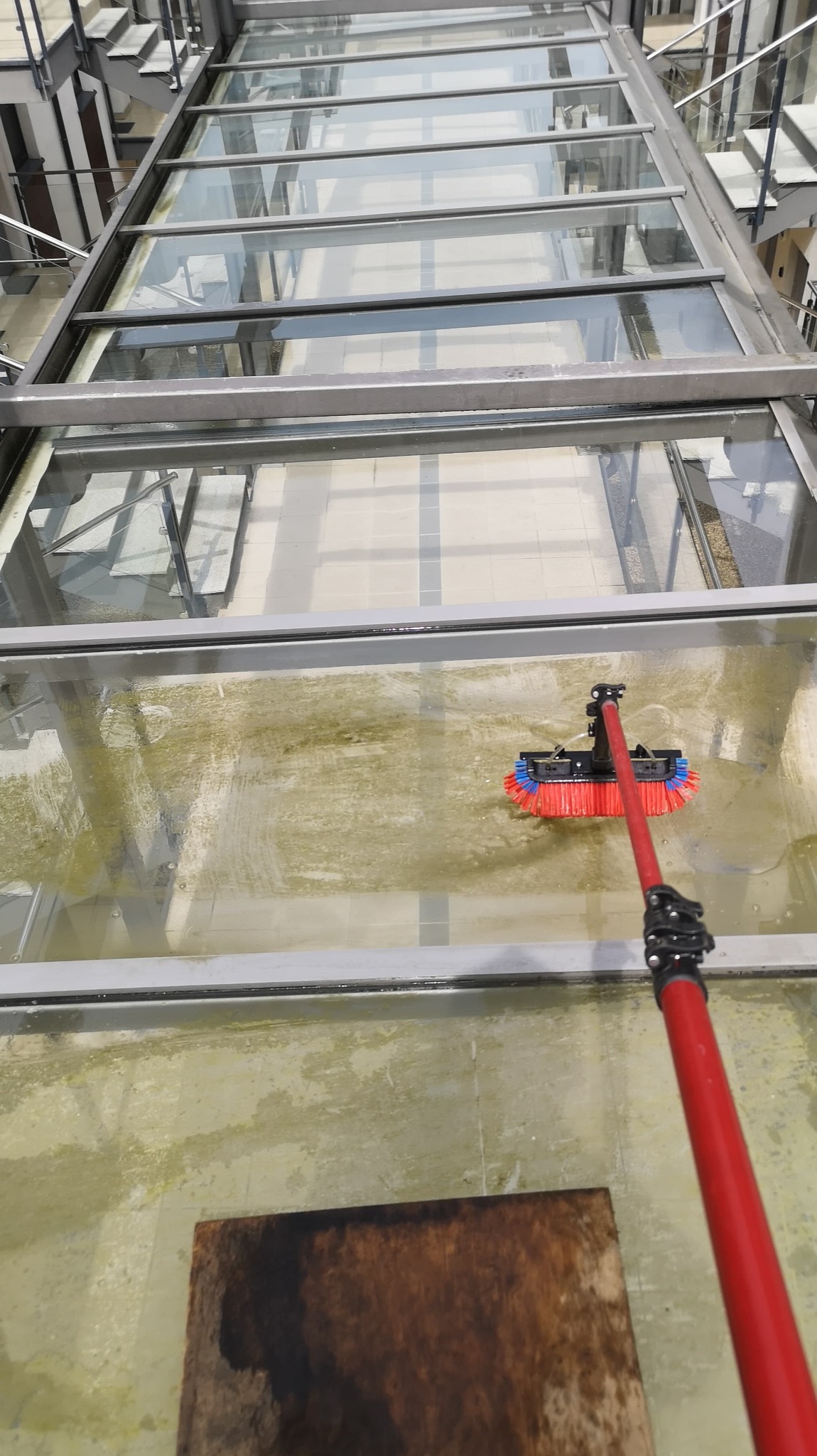 Czyszczenie szklanego dachu z zanieczyszczeń za pomocą szczotki na długim, czerwonym kiju. Widok z góry na brudną i czyszczoną powierzchnię.