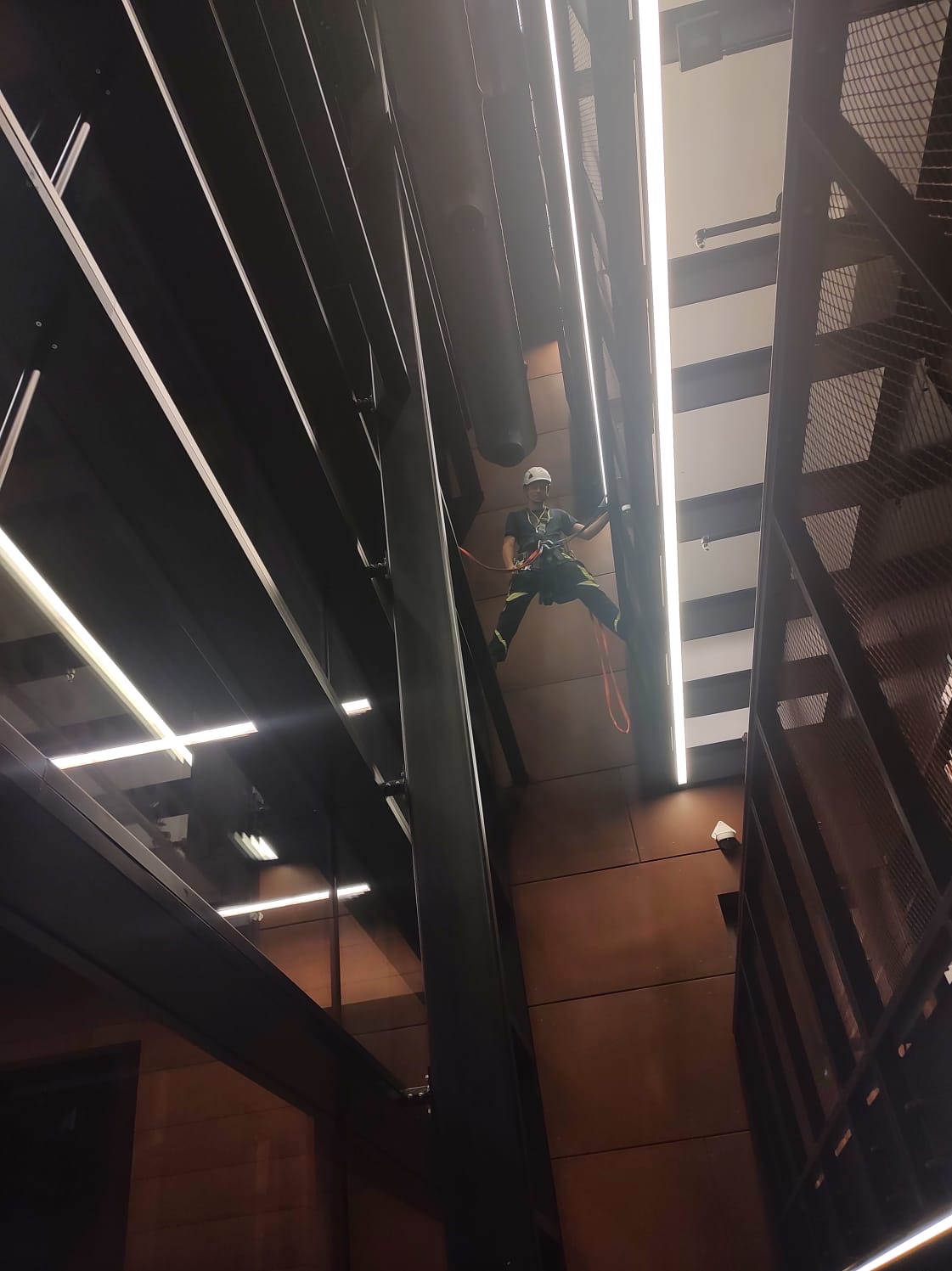 Pracownik w uprzęży, kasku, na wysokości, wewnątrz budynku z metalową konstrukcją i oświetleniem liniowym. Praca na wysokości w przestrzeni komercyjnej.