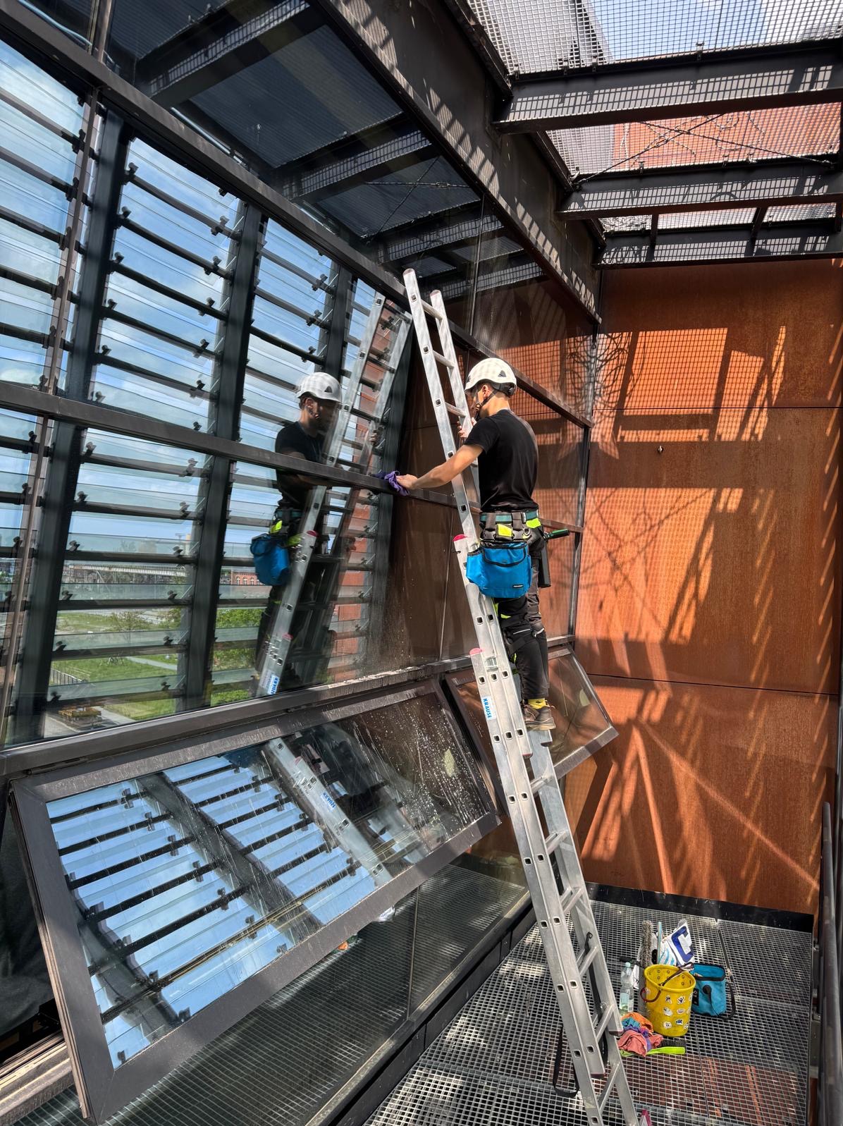Pracownik w uprzęży myje okna na wysokości, stojąc na drabinie. Widoczne refleksy nieba i otoczenia w czyszczonych oknach. Budynek o rdzawym kolorze.