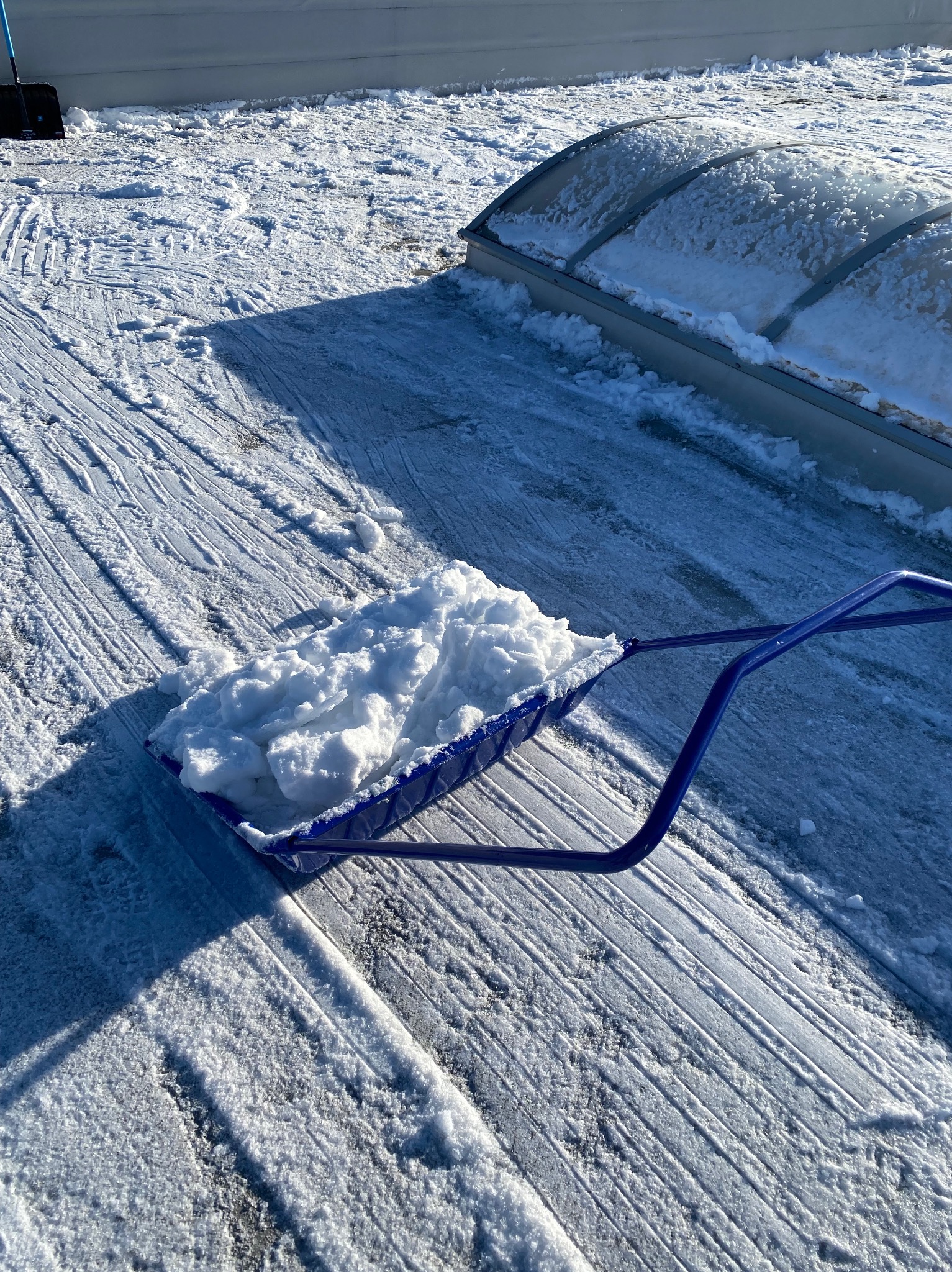 Niebieska łopata pełna śniegu na odśnieżonym dachu z świetlikiem. Widoczne ślady po odśnieżaniu.