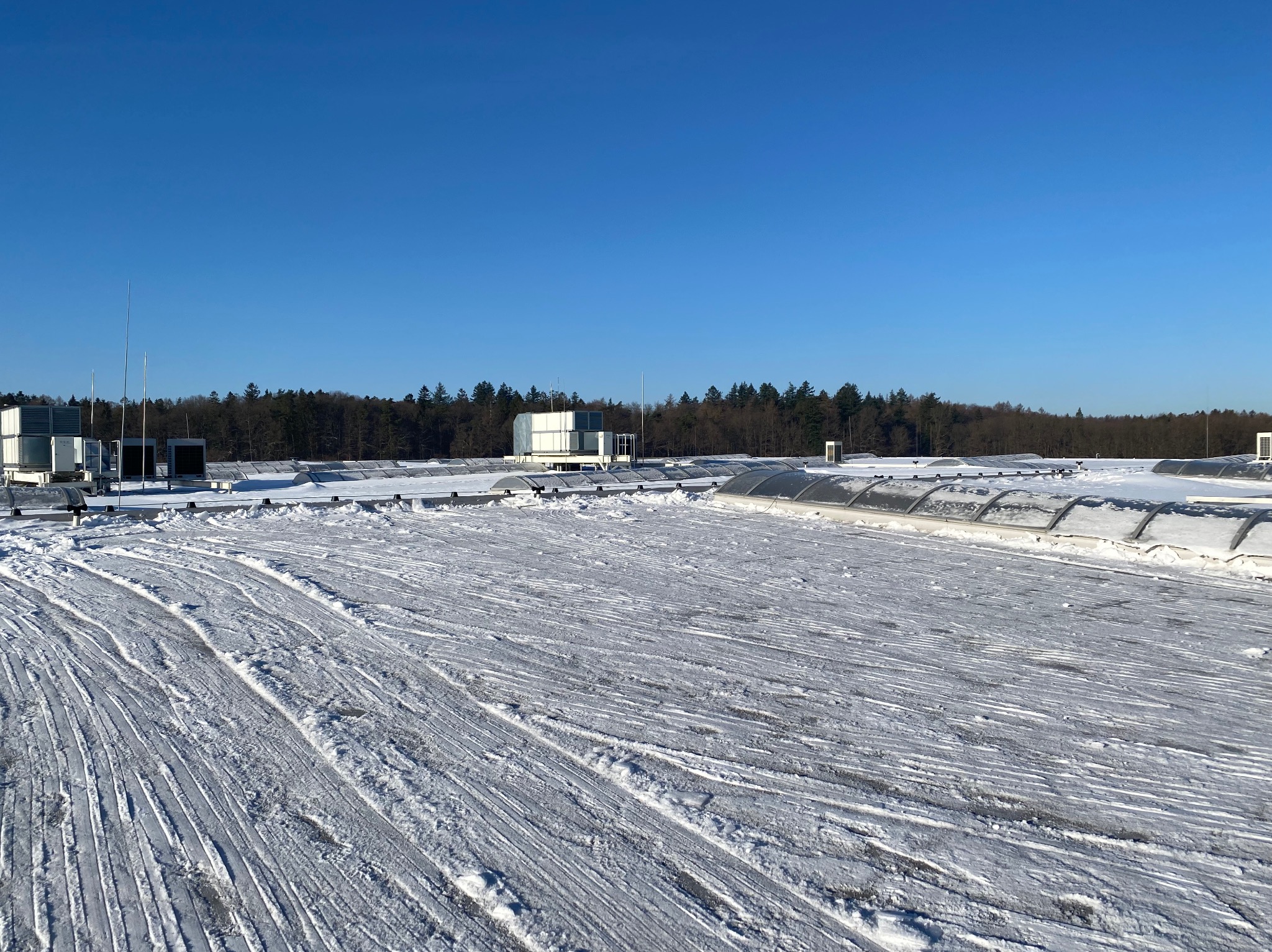 Śnieżny dach przemysłowy z widocznymi śladami odśnieżania, urządzenia wentylacyjne i las w tle, błękitne niebo. Widok z góry, ostre światło słoneczne.