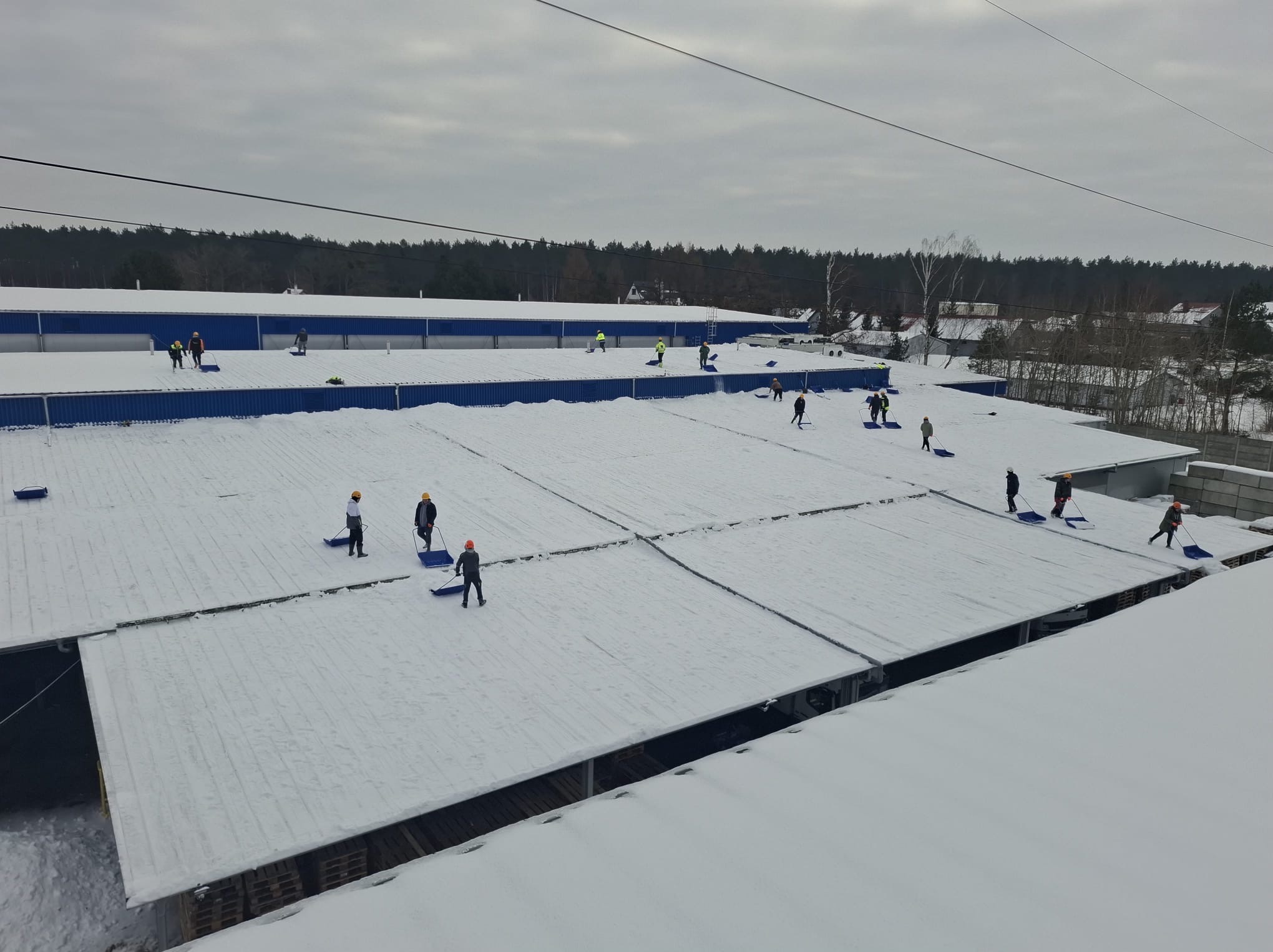 Ekipa w kaskach odśnieża rozległy, płaski dach budynku przemysłowego. Widok z góry ukazuje pracowników z niebieskimi łopatami na tle białej pokrywy śnieżnej i błękitnych krawędzi dachu.