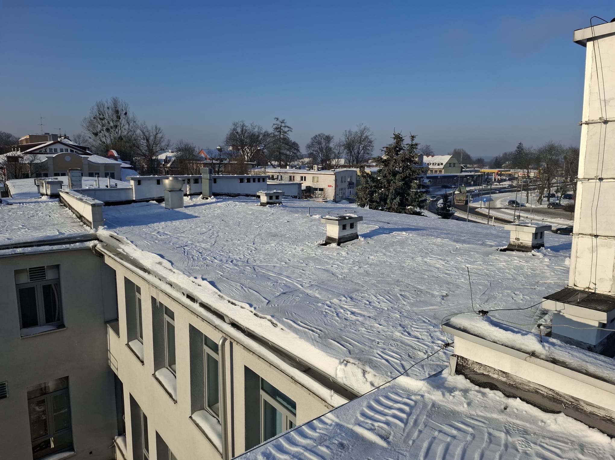 Śnieżny dach budynku z kominami w zimowy dzień, widok z góry. Ślady na śniegu, błękitne niebo i zabudowa w tle. Widoczne okna budynku.