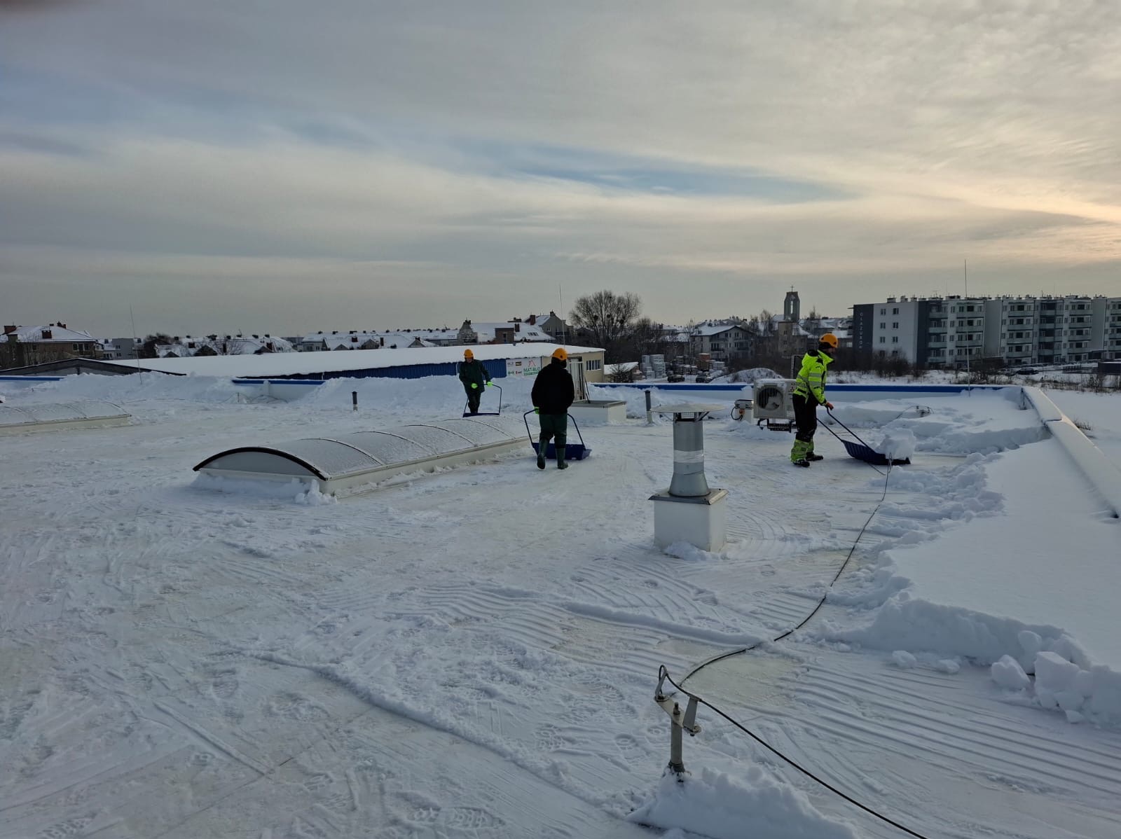 Pracownicy w kaskach odśnieżają płaski dach budynku za pomocą łopat. Widoczne odśnieżone fragmenty dachu i budynki w tle.