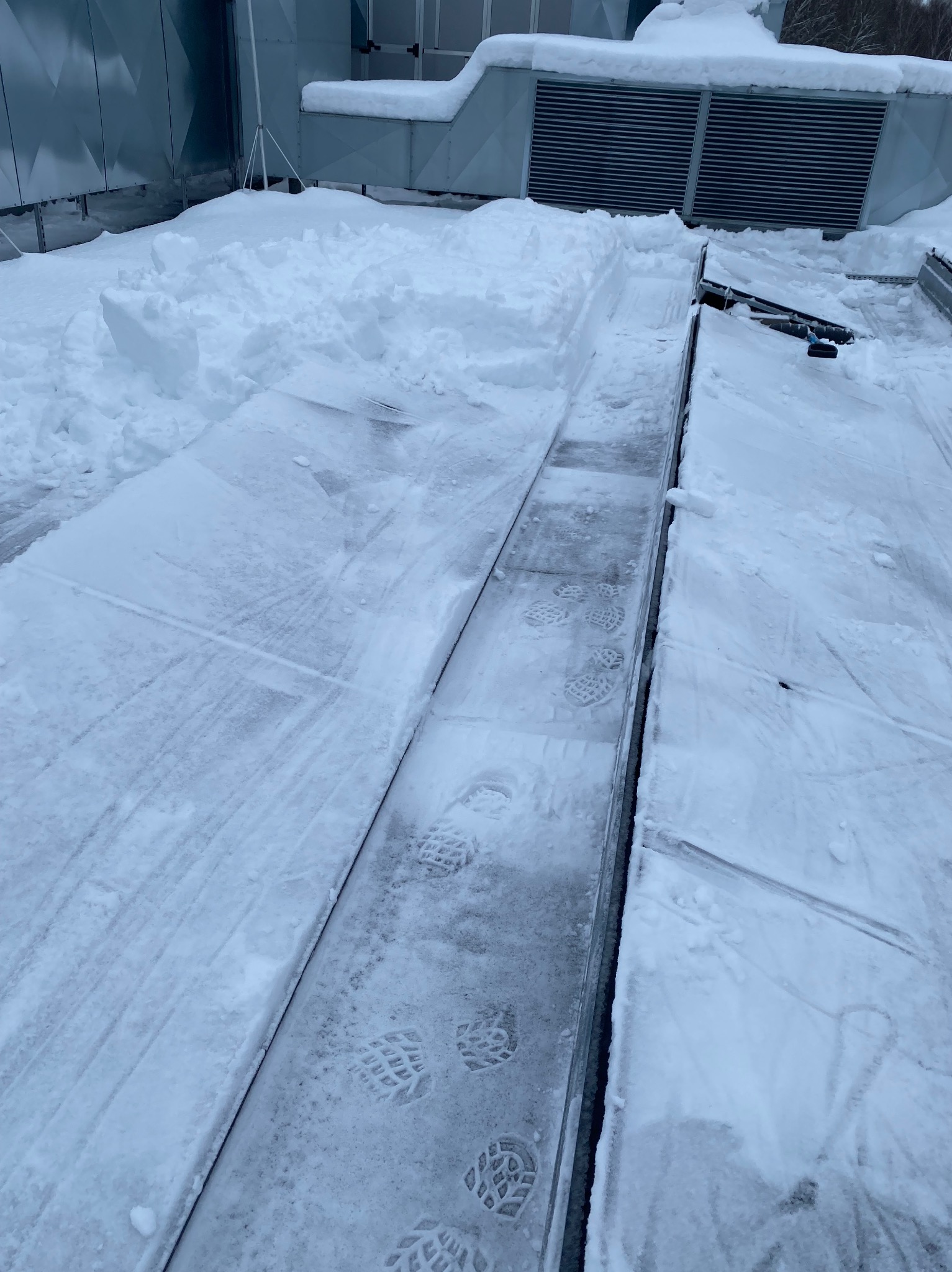 Odśnieżony fragment dachu z widocznymi śladami butów w śniegu, obok leżące pryzmy śniegu. W tle instalacja wentylacyjna pokryta śniegiem.