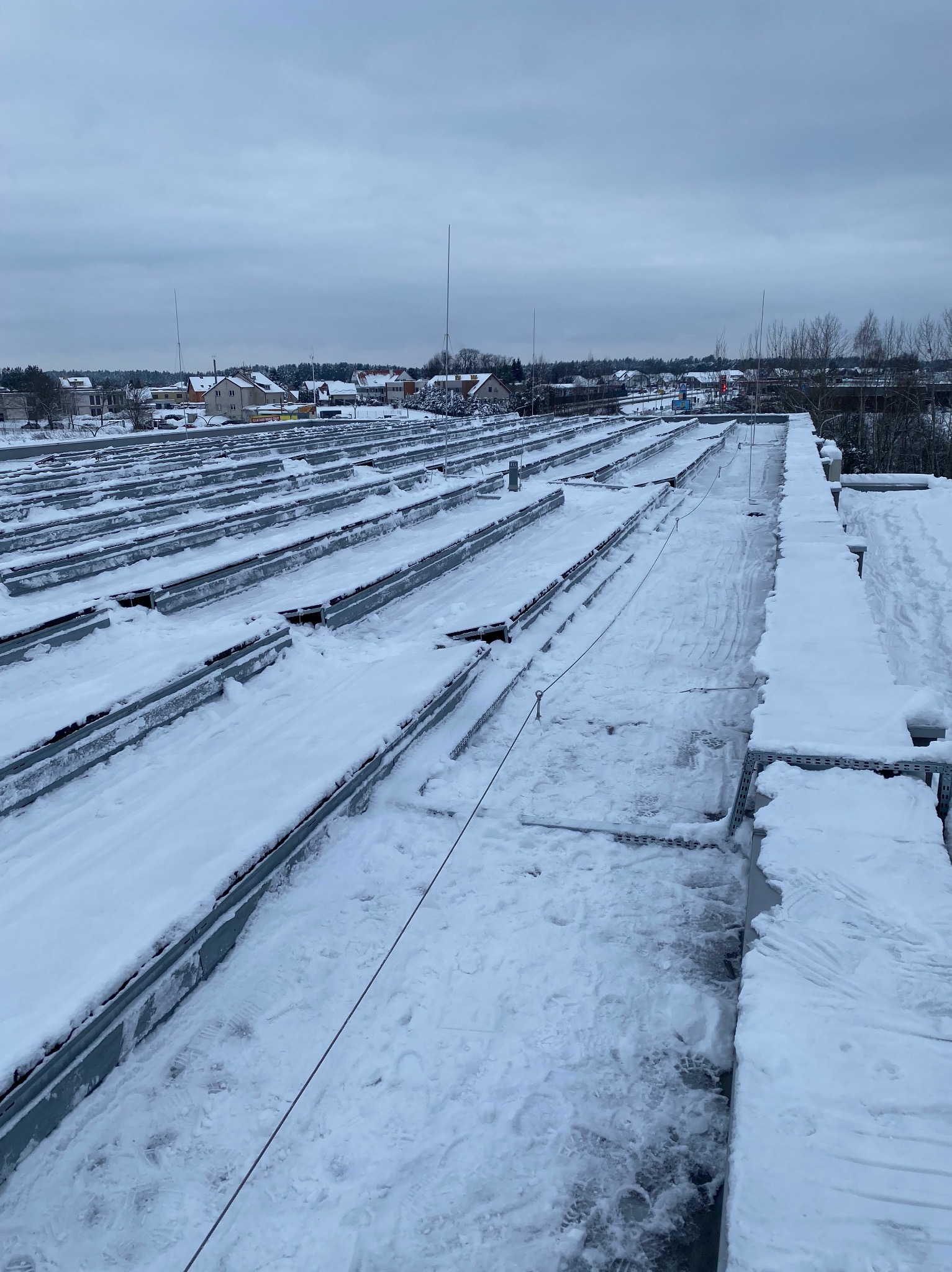 Śnieg na dachu budynku z widocznymi elementami konstrukcyjnymi i śladami stóp, w tle zabudowa miejska pod pochmurnym niebem.