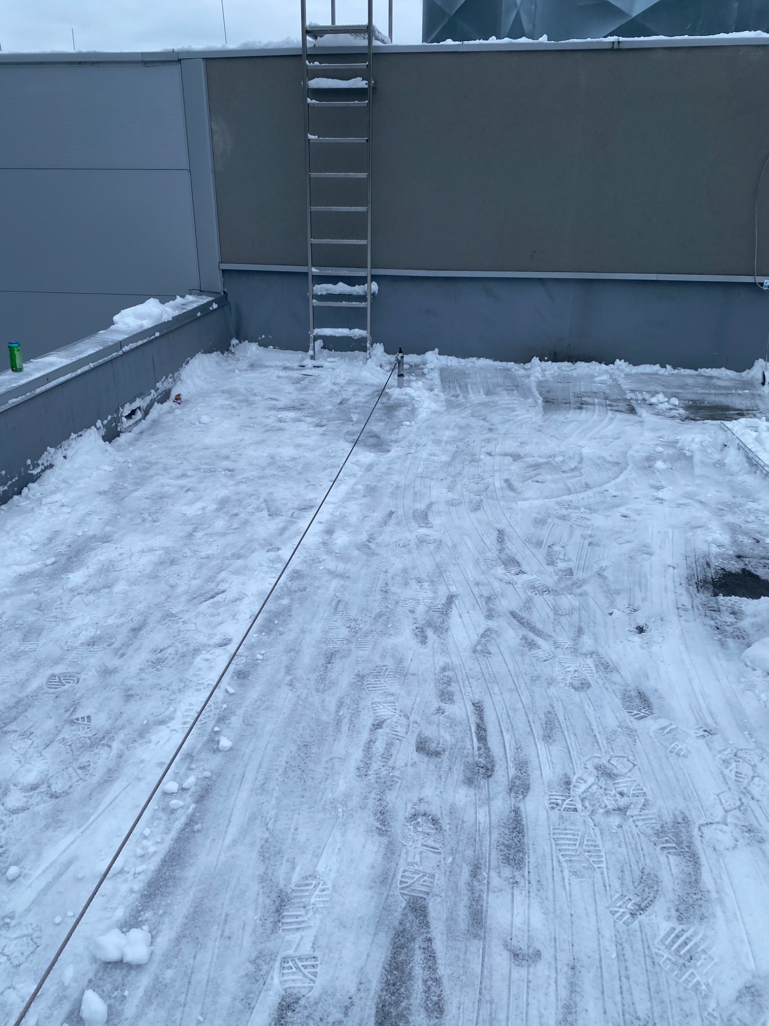 Śnieg na dachu budynku po odśnieżaniu, widoczne ślady butów i lina zabezpieczająca. Metalowa drabina oparta o ścianę. Szary budynek w tle.