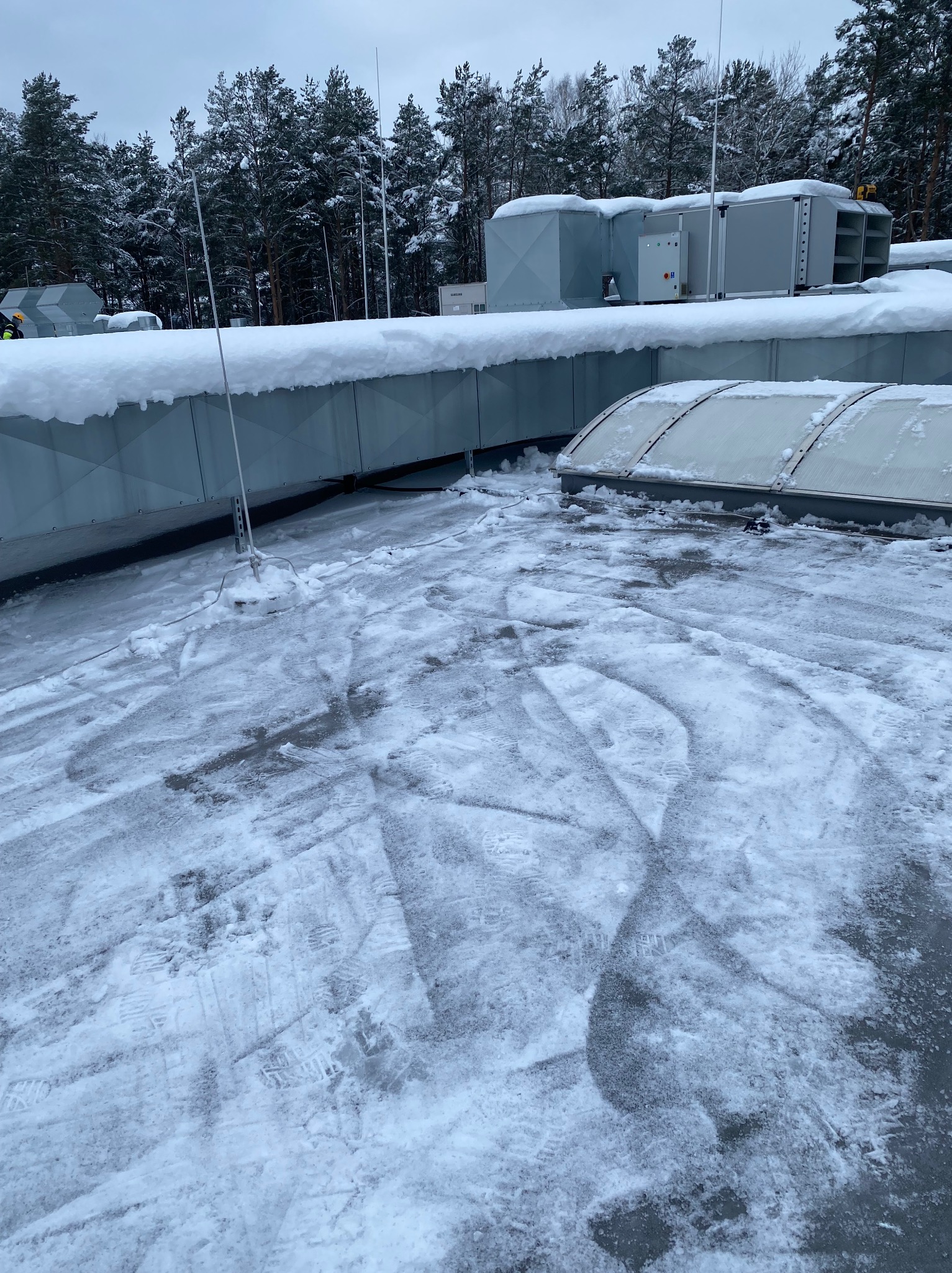 Zaśnieżony dach budynku z instalacjami wentylacyjnymi w Elblągu. Widoczne ślady stóp na śniegu i drzewa w tle.