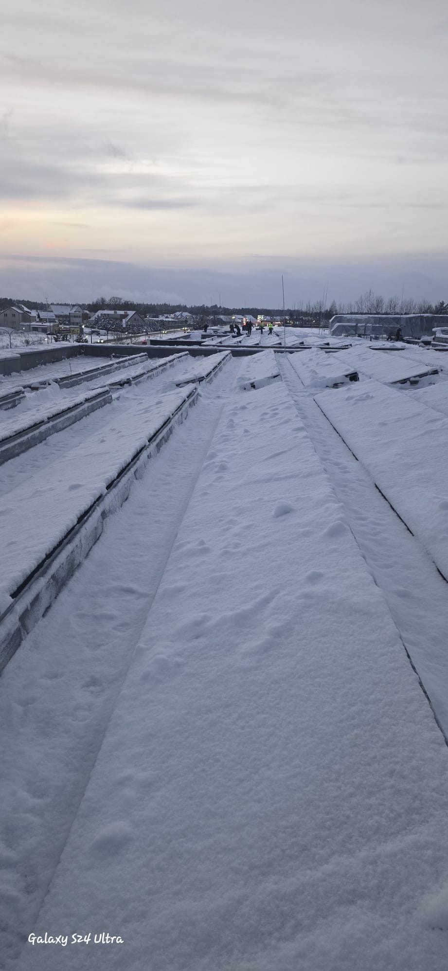 Śnieg na dachu budynku z widocznymi elementami konstrukcyjnymi i pracownikami w oddali, sfotografowany w pochmurny dzień.