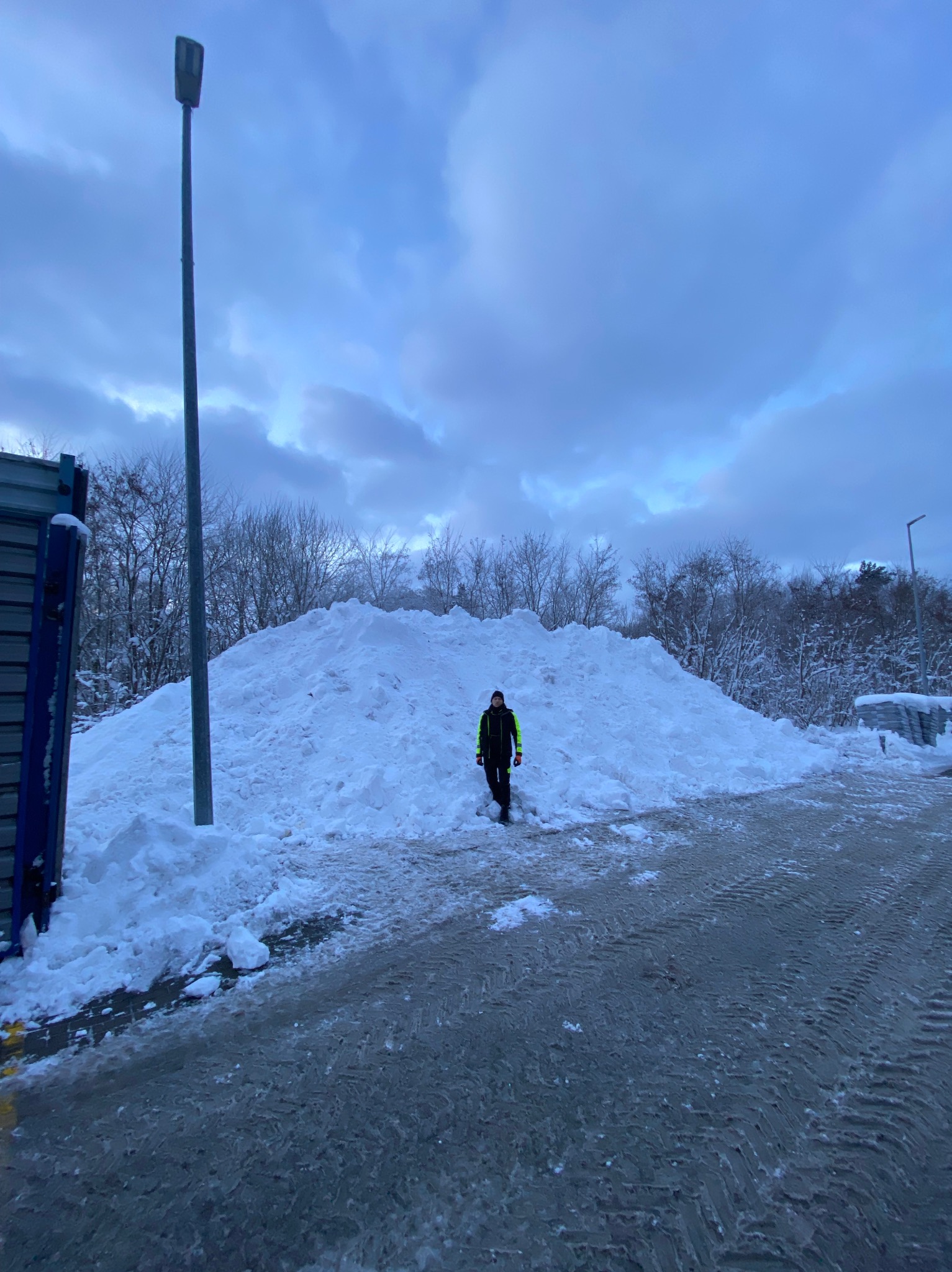 Mężczyzna w odblaskowej kurtce stoi na tle ogromnej pryzmy śniegu, prawdopodobnie po odśnieżaniu. Widoczne ślady opon na oblodzonej nawierzchni i pochmurne niebo.