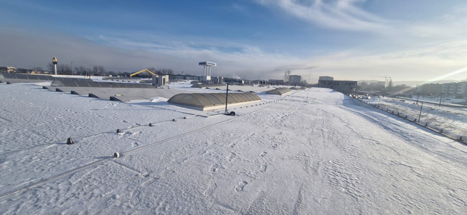 Śnieżny dach hipermarketu z świetlikami i śladami stóp, widok z góry w słoneczny dzień. W tle zabudowa miejska i błękitne niebo.