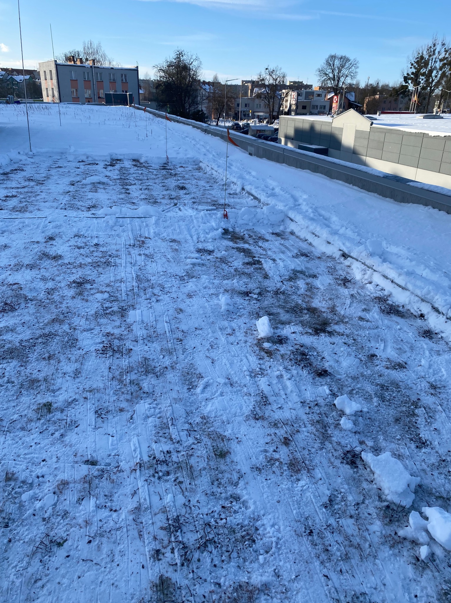 Śnieg na płaskim dachu budynku z widocznymi śladami odśnieżania i pomarańczowymi znacznikami. W tle zabudowa miejska i błękitne niebo.