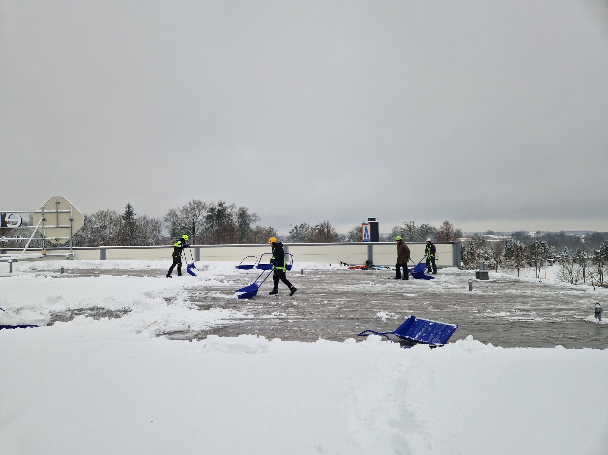 Pracownicy w odblaskowych ubraniach odśnieżają płaski dach budynku za pomocą niebieskich odśnieżarek na kółkach. Widoczny krajobraz zaśnieżonego terenu.