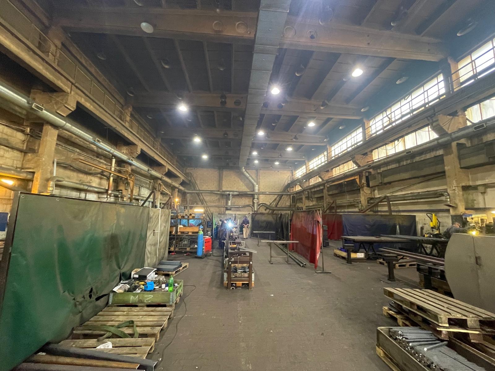 Wnętrze hali produkcyjnej z widocznymi stanowiskami pracy, narzędziami i materiałami. Widoczne okna w górnej części hali i oświetlenie sufitowe.