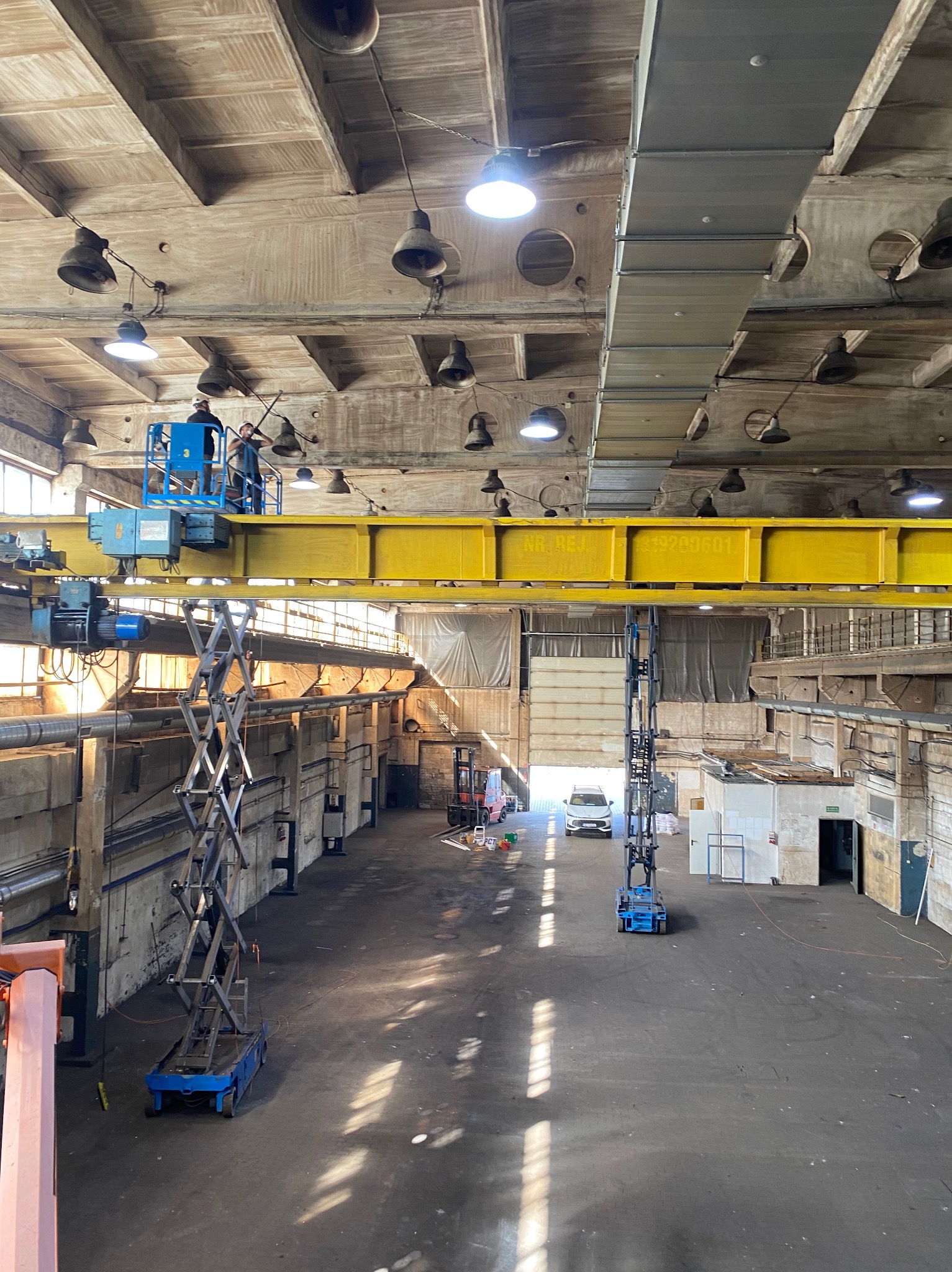 Pracownicy na podnośniku koszowym czyszczą sufit hali przemysłowej z licznymi lampami. Widoczny dźwig suwnicowy i wózek widłowy w tle.