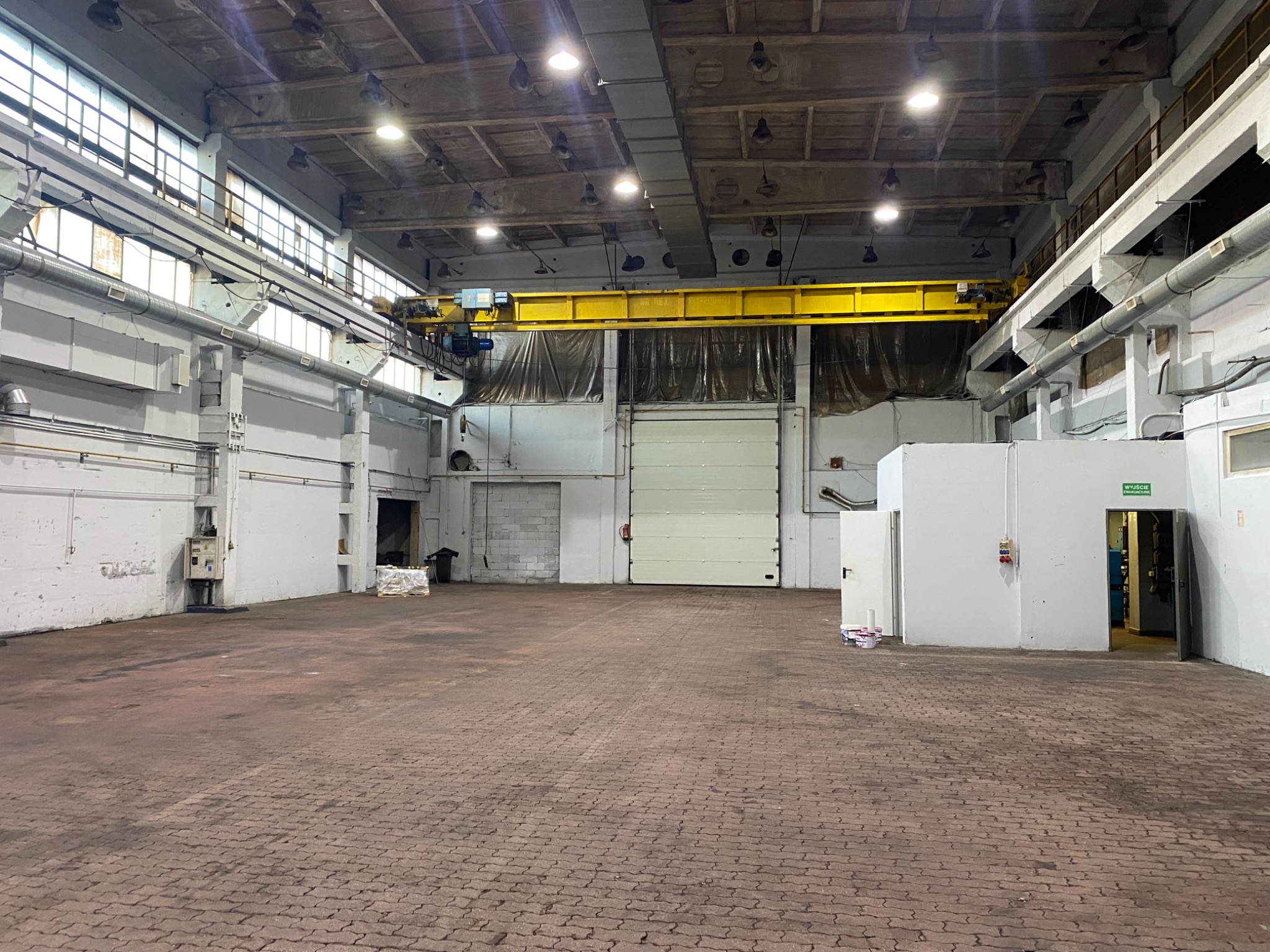 Przestronne wnętrze hali przemysłowej z suwnicą, ceglaną posadzką i białymi ścianami. Widoczne okna, oświetlenie i brama wjazdowa. Hala przygotowana do prac porządkowych.