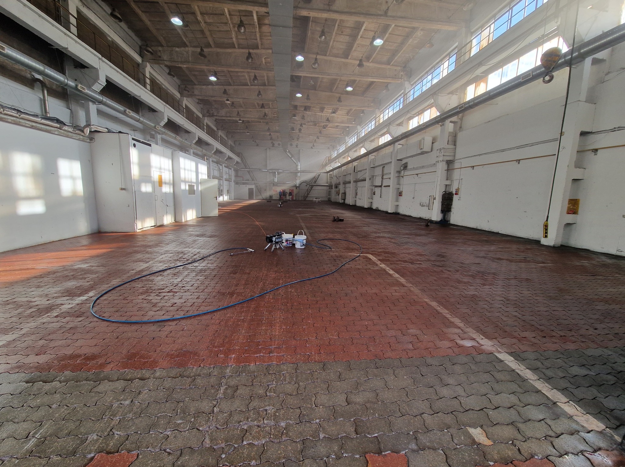 Wnętrze hali przemysłowej z częściowo umytą podłogą z kostki brukowej. Widoczny sprzęt do mycia ciśnieniowego i węże. W tle osoby w oddali.