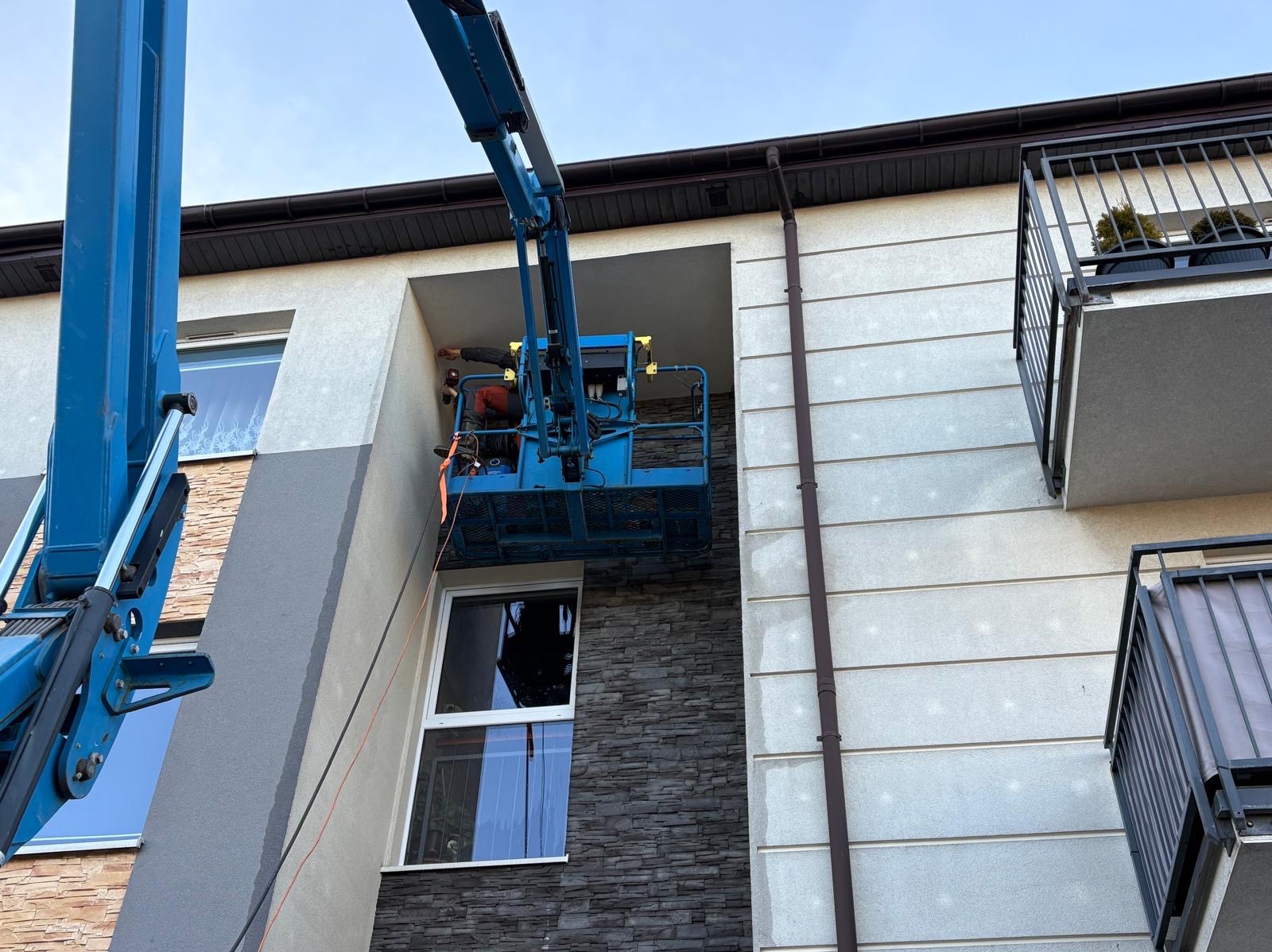 Pracownik w podnośniku koszowym, wykonujący prace na elewacji budynku wielorodzinnego z oknami i balkonami. Widoczne fragmenty elewacji w odcieniach szarości i beżu.