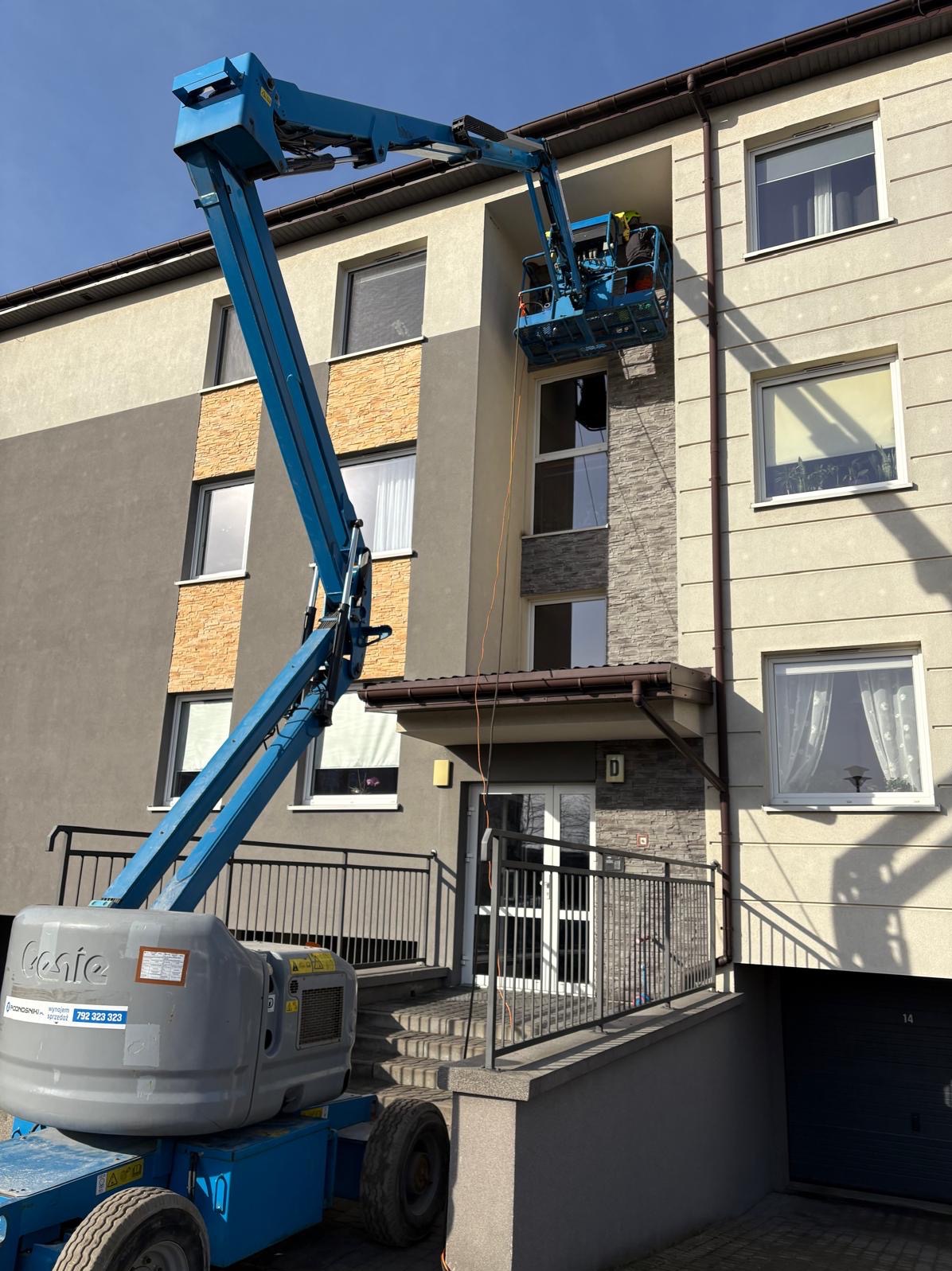 Pracownik w koszu podnośnika teleskopowego czyści okna na wysokości w Elblągu. Widoczna elewacja budynku z oknami i fragment podnośnika.