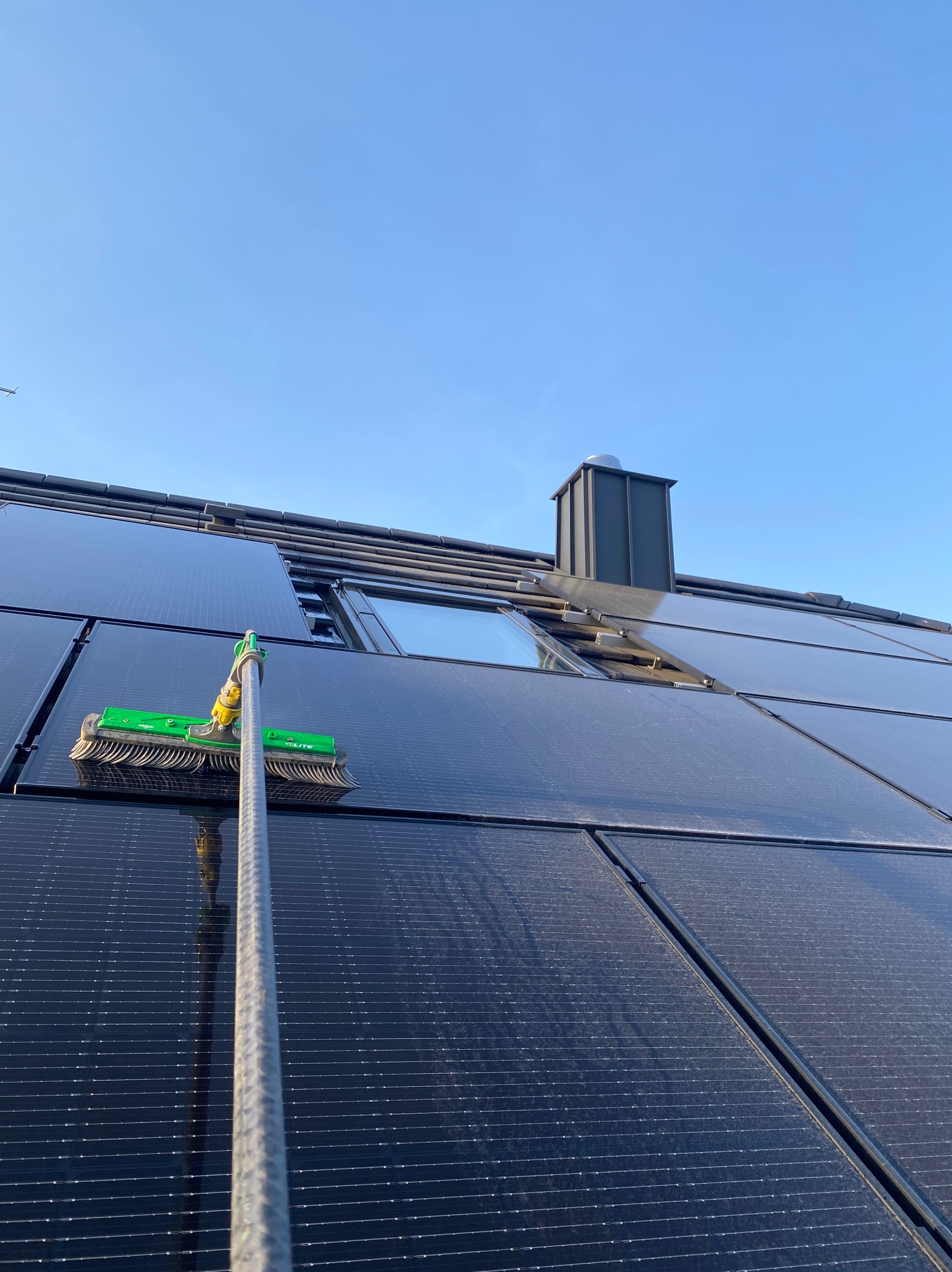 Szczotka na teleskopowym kiju czyści panele słoneczne na dachu domu. Widoczny komin i okno dachowe. Błękitne niebo w tle. Ujęcie z dołu.